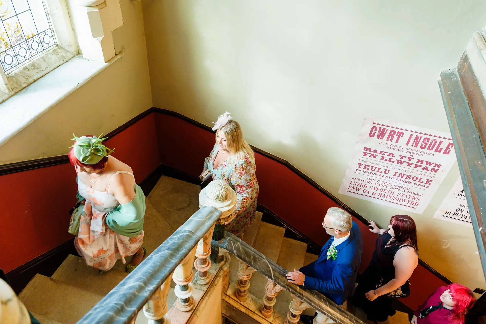 A high-angle shot captures wedding guests dressed in formal attire ascending a grand indoor staircase with a marble handrail.