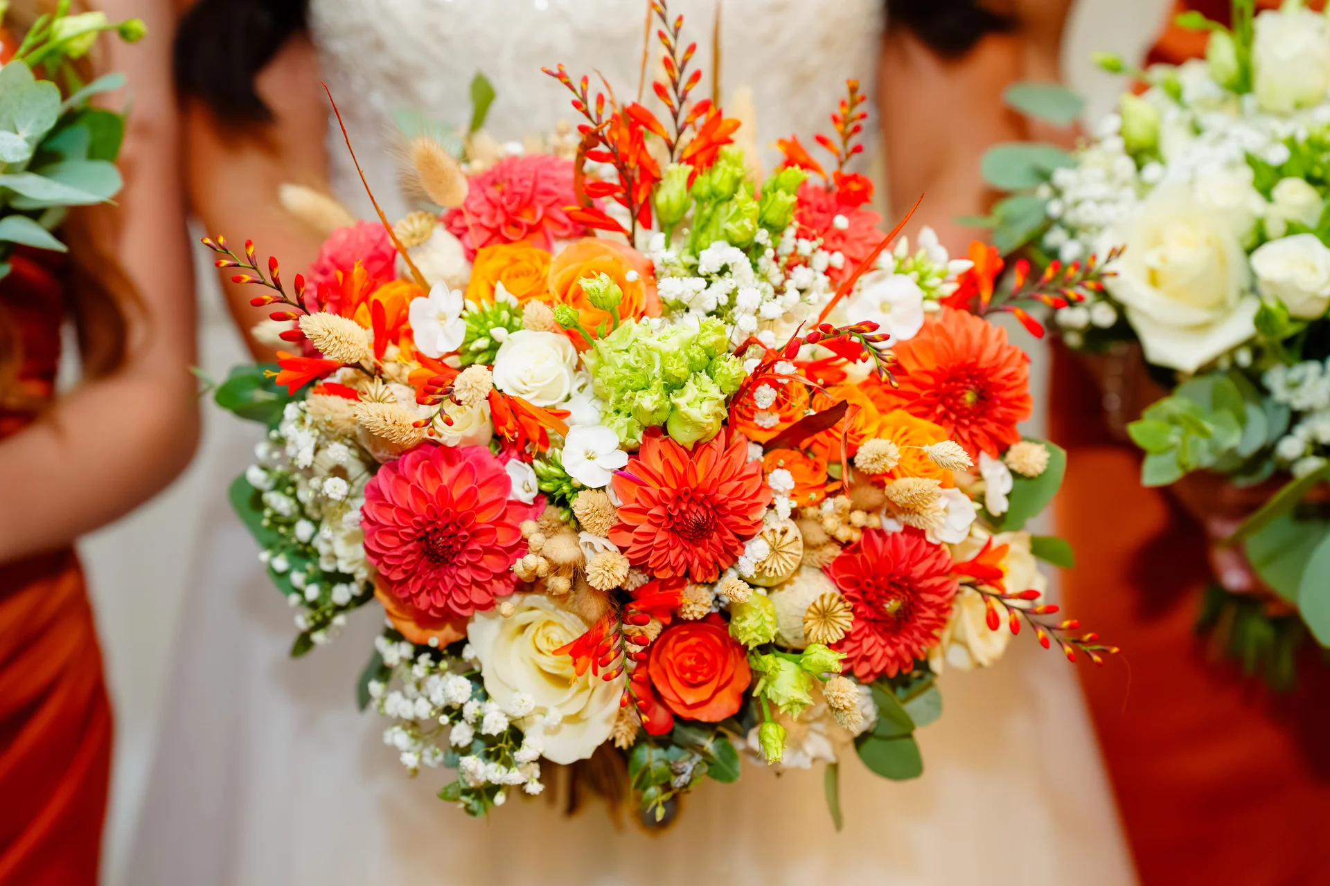 A vibrant hand-tied bridal bouquet featuring burnt orange and red dahlias, white roses, and delicate filler flowers held against a white gown.