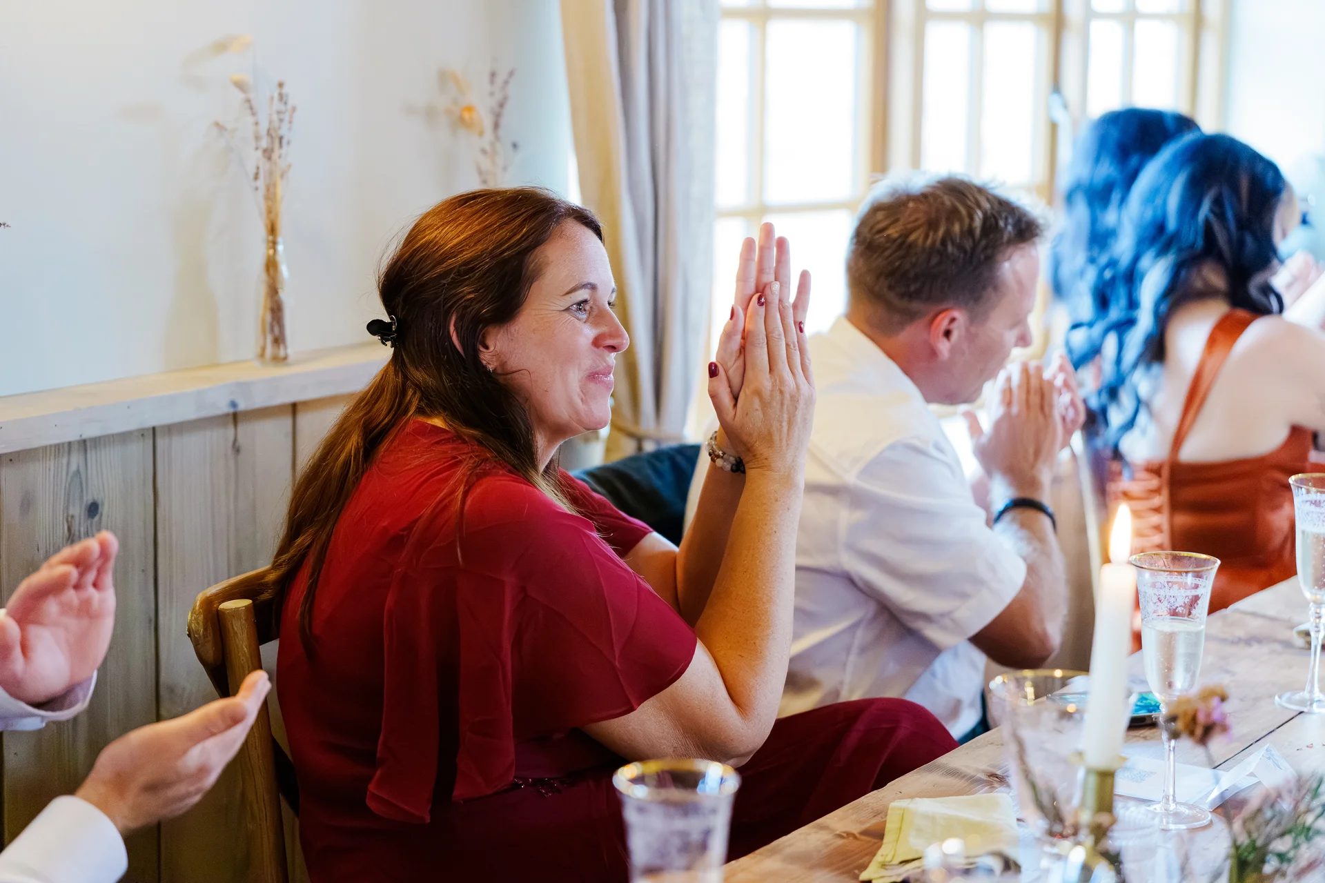 Wedding guests sit at a rustic wooden banquet table decorated with candles, clapping and smiling happily during the wedding speeches.