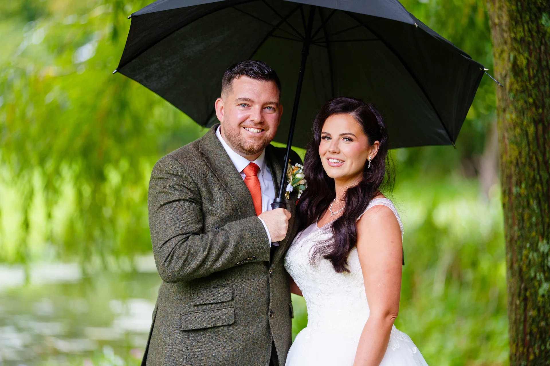 A smiling bride and groom pose together outdoors under a large black umbrella amidst lush green trees.