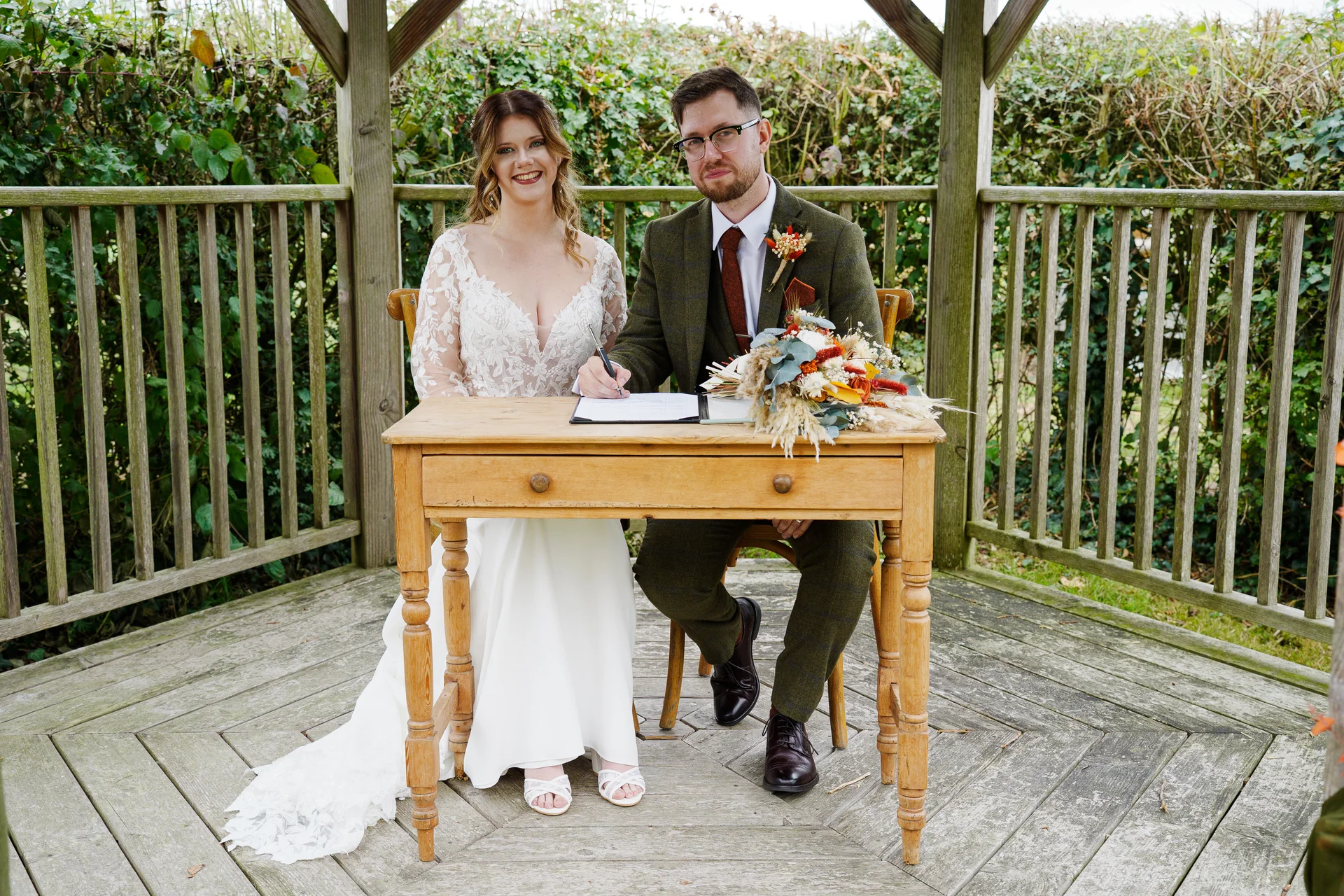 A smiling bride and groom sit together inside a wooden gazebo while signing the wedding register behind a vintage wooden desk.