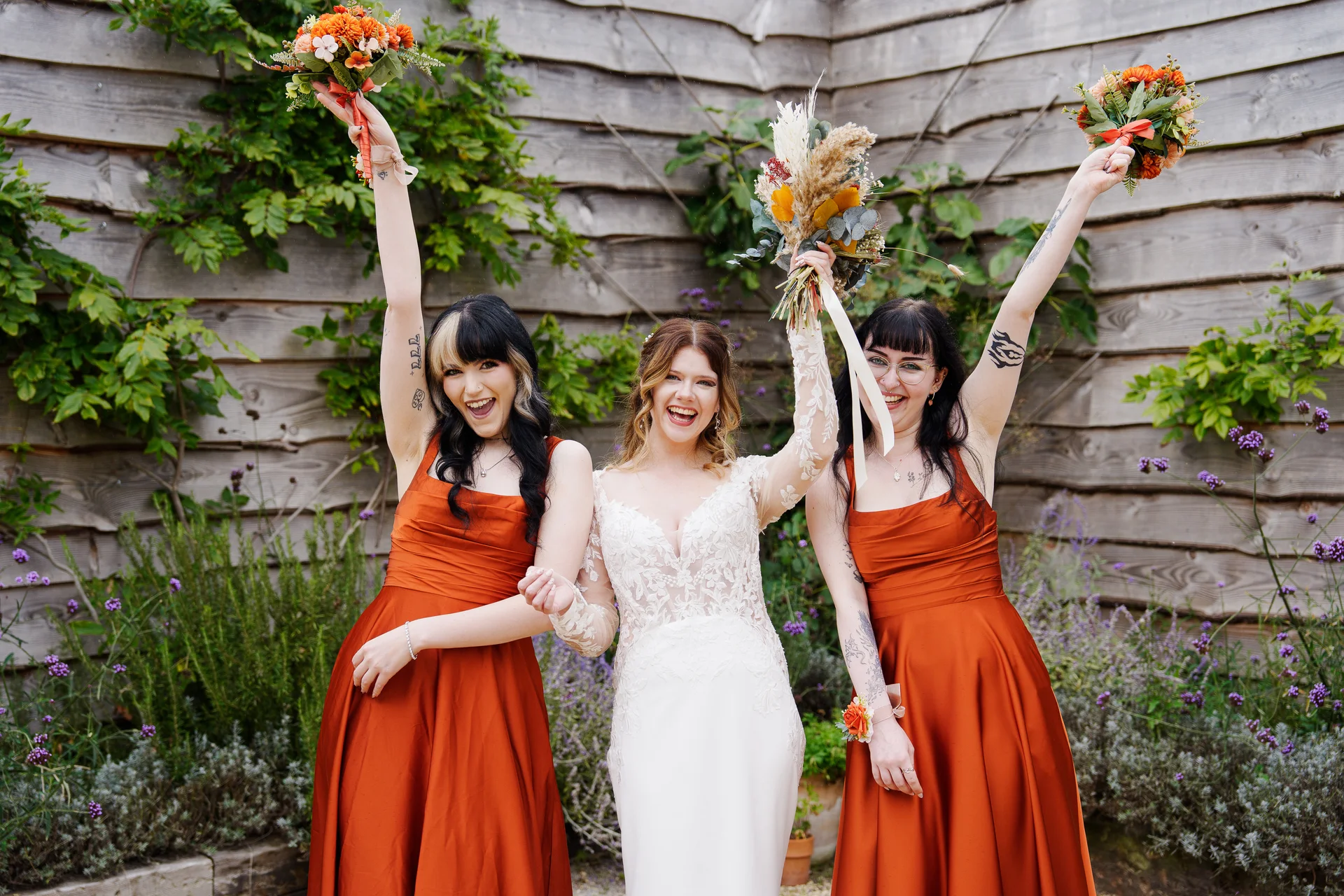 A joyful bride and her bridesmaids in satin burnt orange dresses raise their bouquets in celebration against a rustic wooden wall.