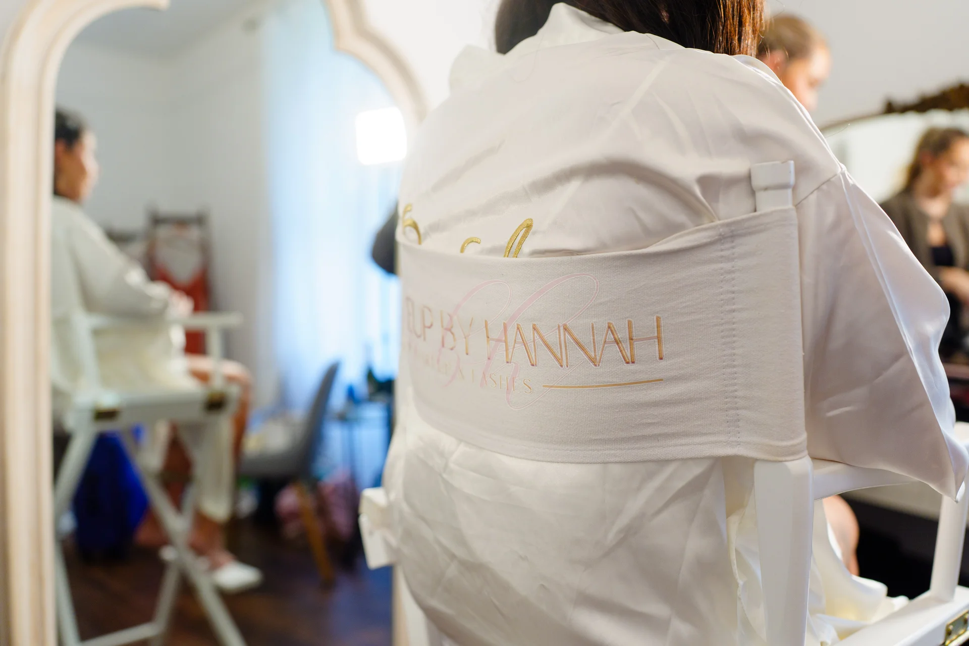 A bride sits in a white makeup chair wearing a bridal robe while facing a mirror during morning bridal preparations.