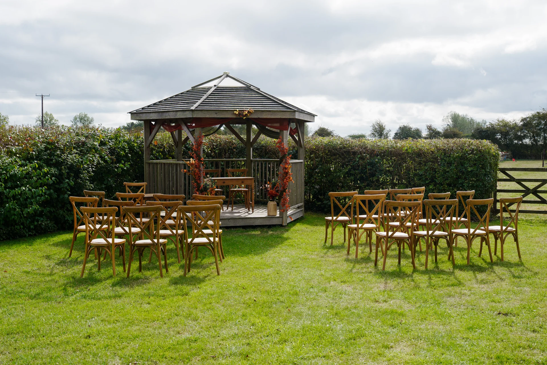 An outdoor wedding ceremony setup features a wooden gazebo decorated with autumnal florals and arranged wooden cross back chairs on a lush green lawn.