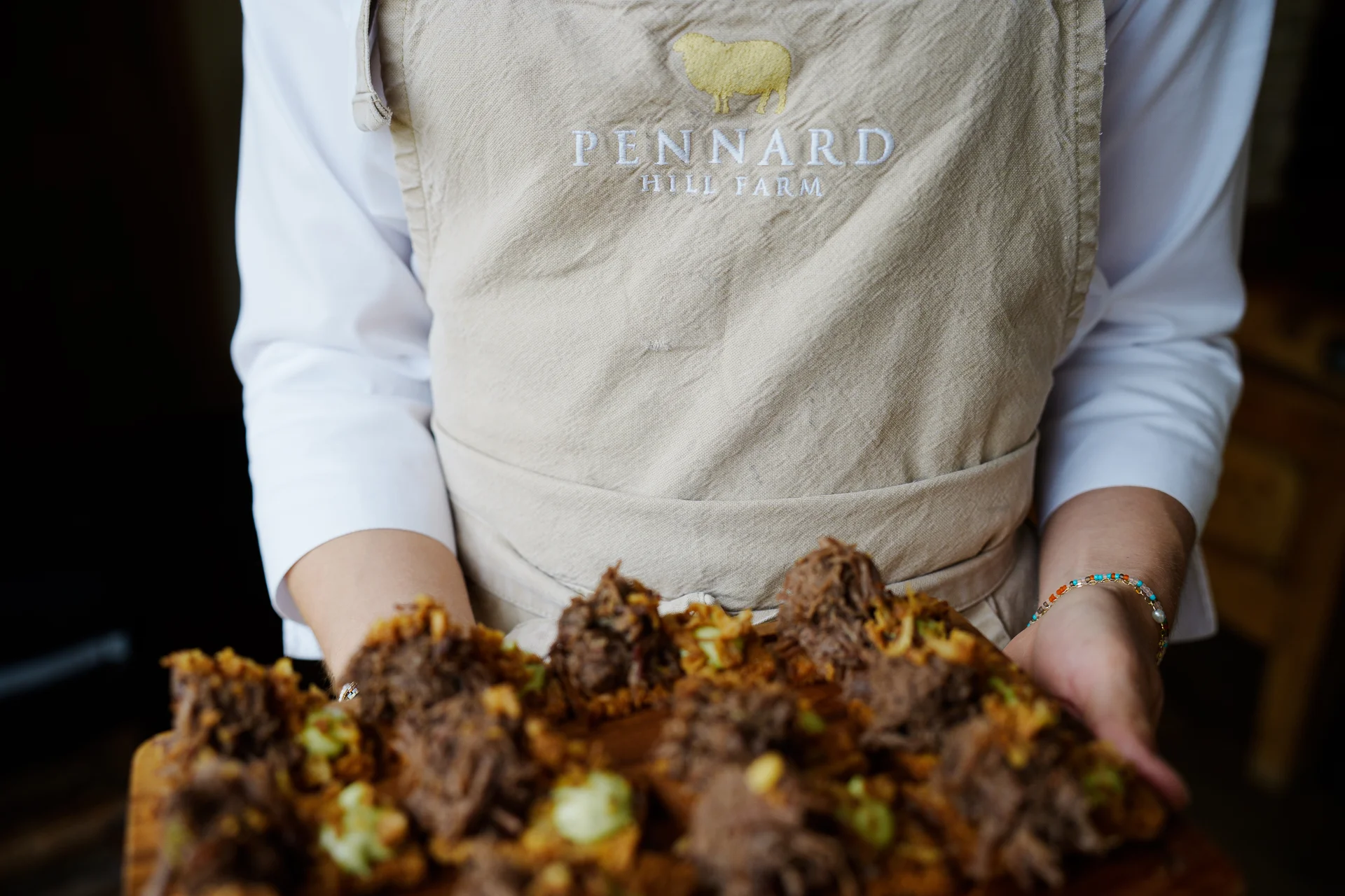 A member of catering staff wearing a branded linen apron holds a wooden board laden with gourmet canapés at a farm wedding.