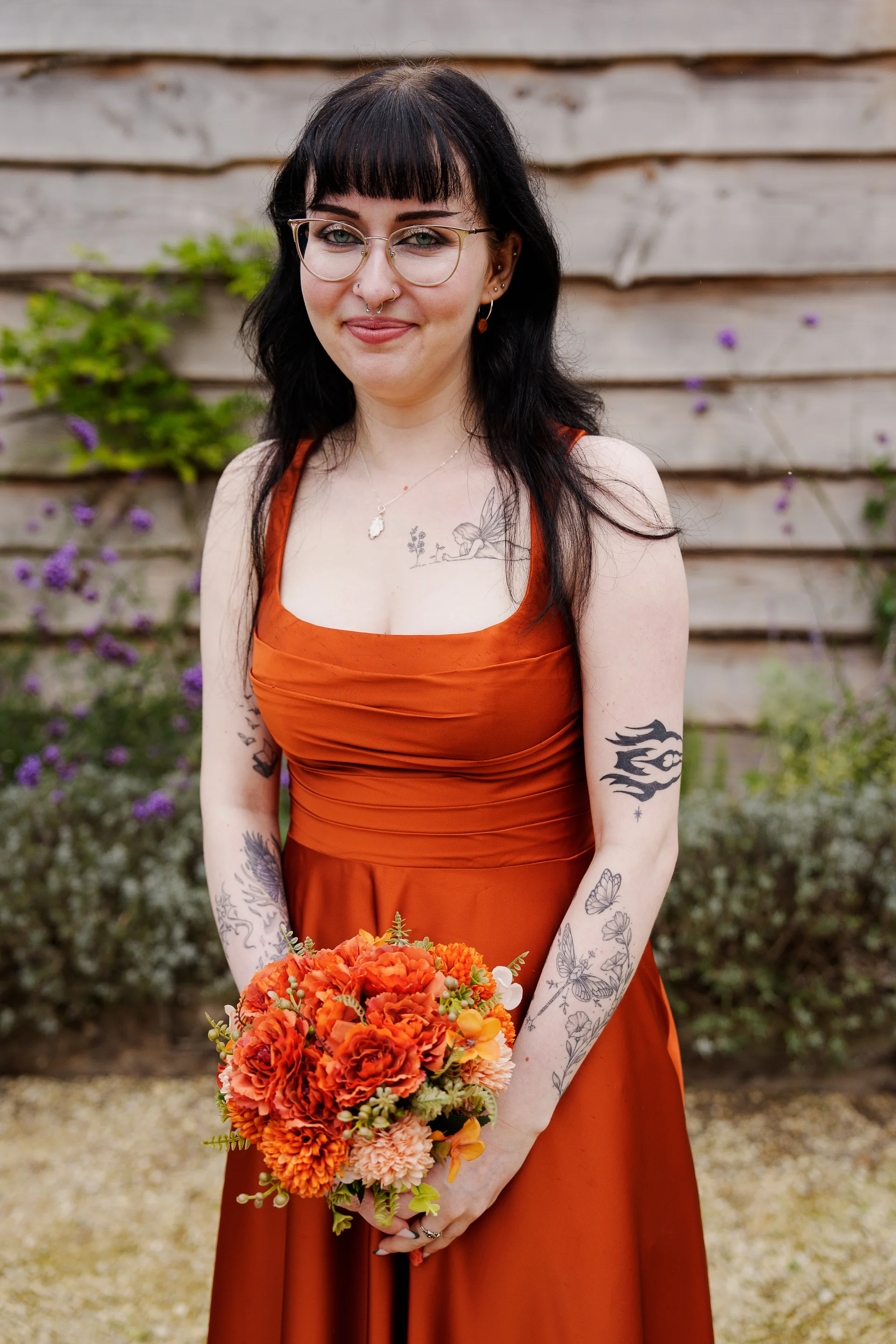 A smiling bride wearing glasses and an orange wedding dress stands in front of a wooden wall, holding a vibrant orange bouquet.