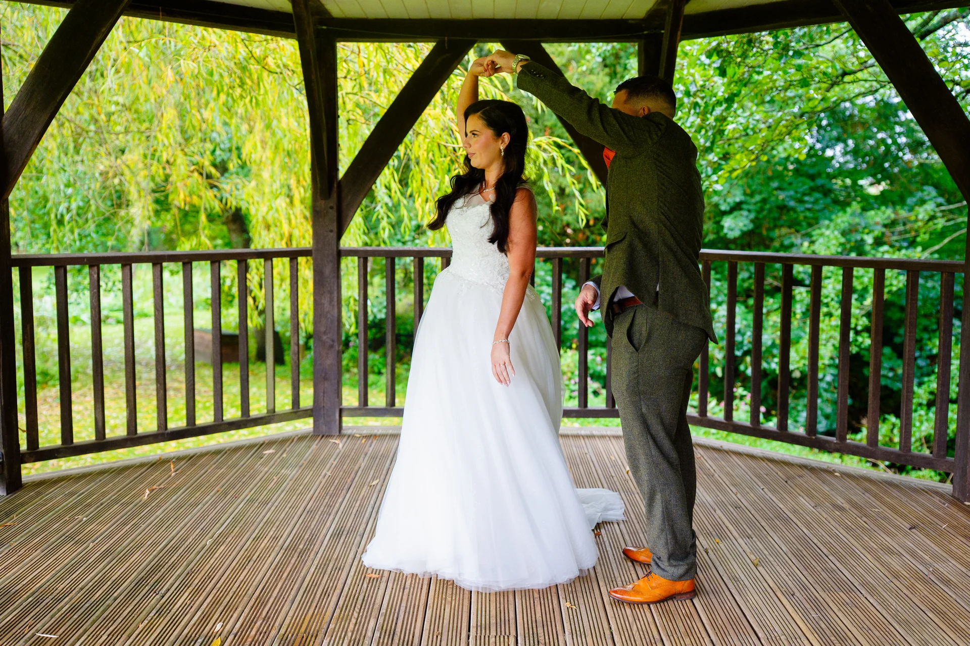 Newlyweds share a romantic dance together inside a wooden bandstand surrounded by lush green foliage and trees.