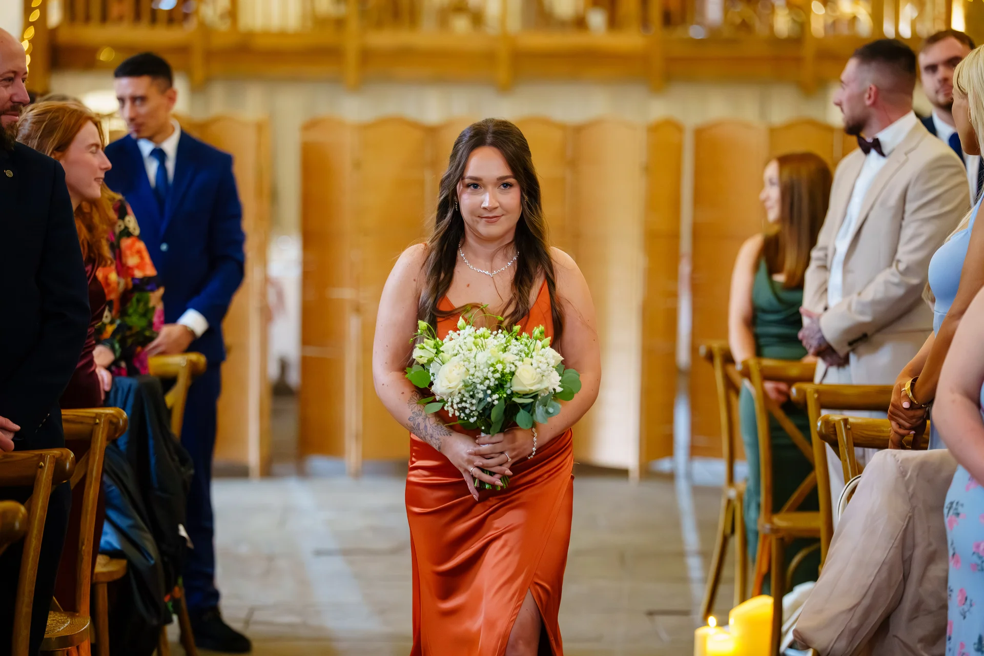 A bridesmaid in a rust-coloured satin slip dress walks down the aisle holding a bouquet of white roses and greenery inside a rustic timber barn.
