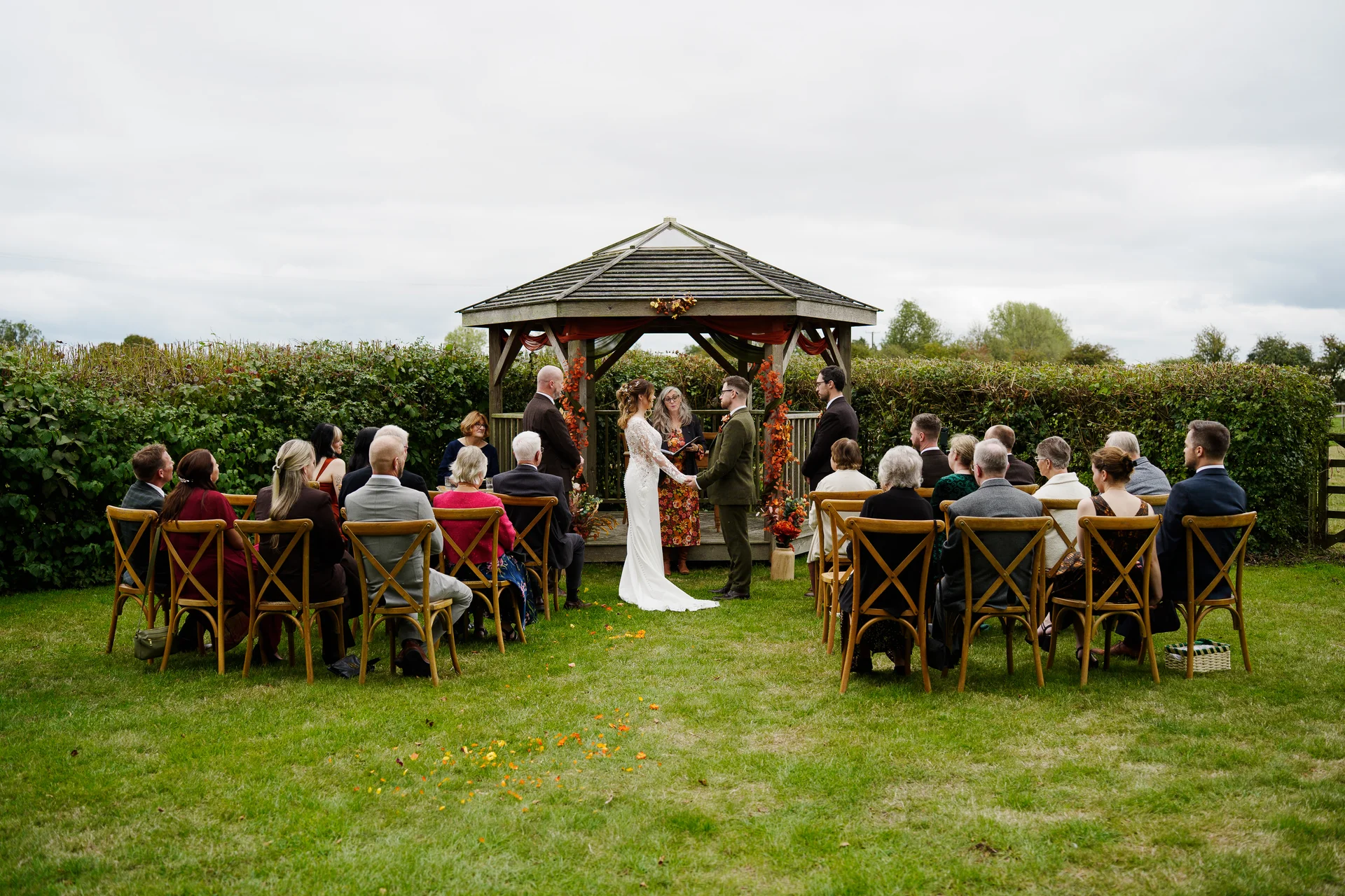 A bride in a long-sleeved gown and groom in a green suit exchange vows during an outdoor celebrant-led ceremony inside a gazebo, surrounded by seated guests on a lush lawn.