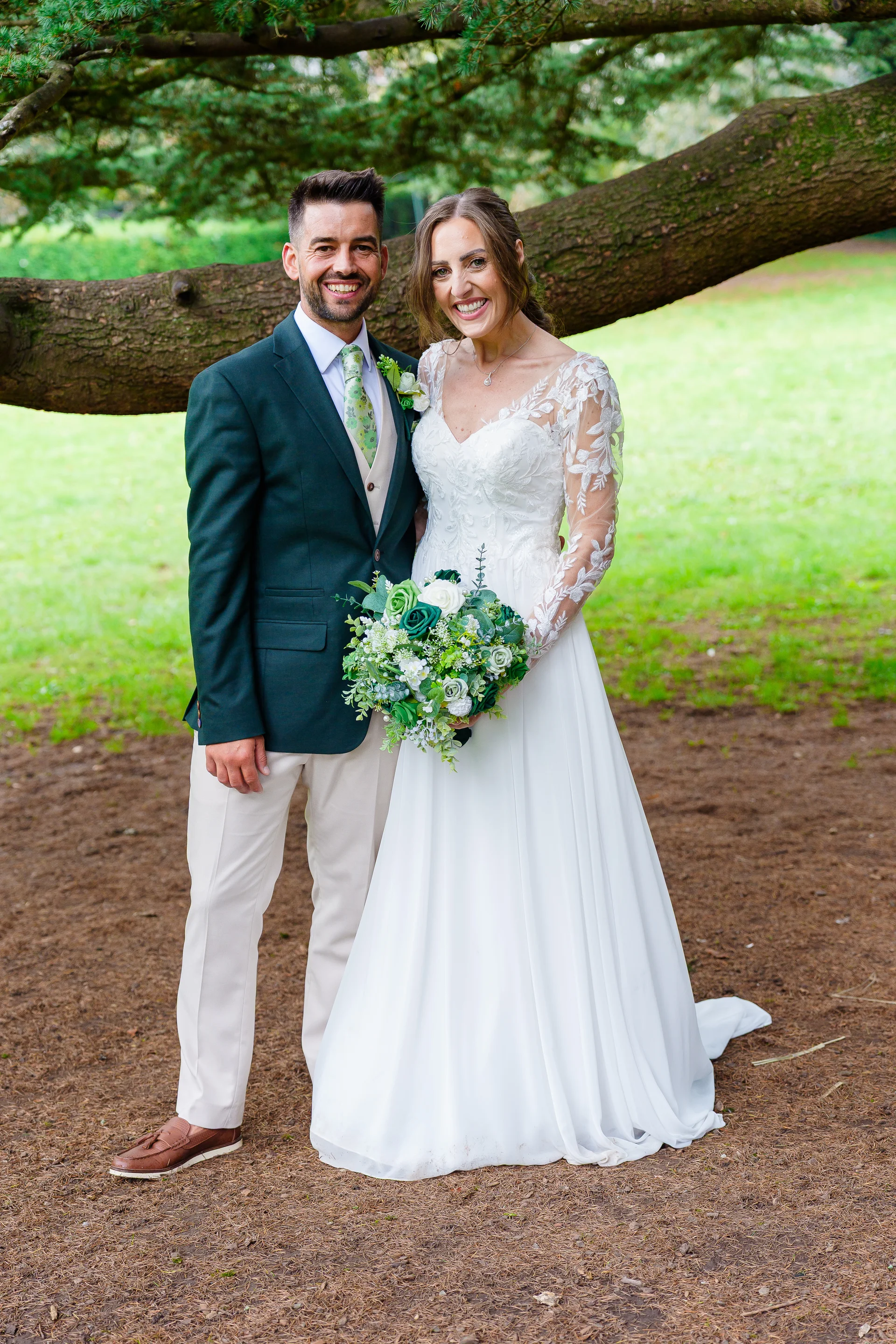 A smiling bride and groom pose together outdoors in a woodland setting beneath a large horizontal tree branch, with the bride holding a lush white and green bouquet.