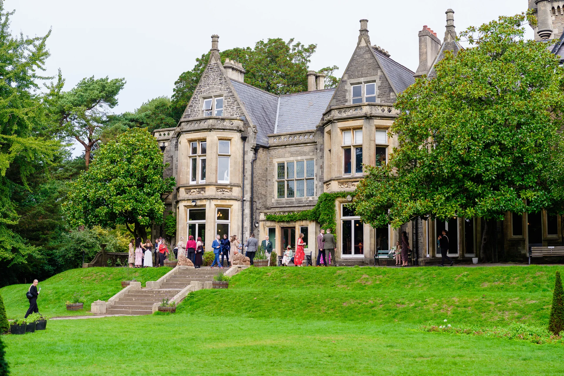 A wide angle documentary shot captures wedding guests mingling and chatting on the sweeping manicured lawn in front of a grand stone country house.