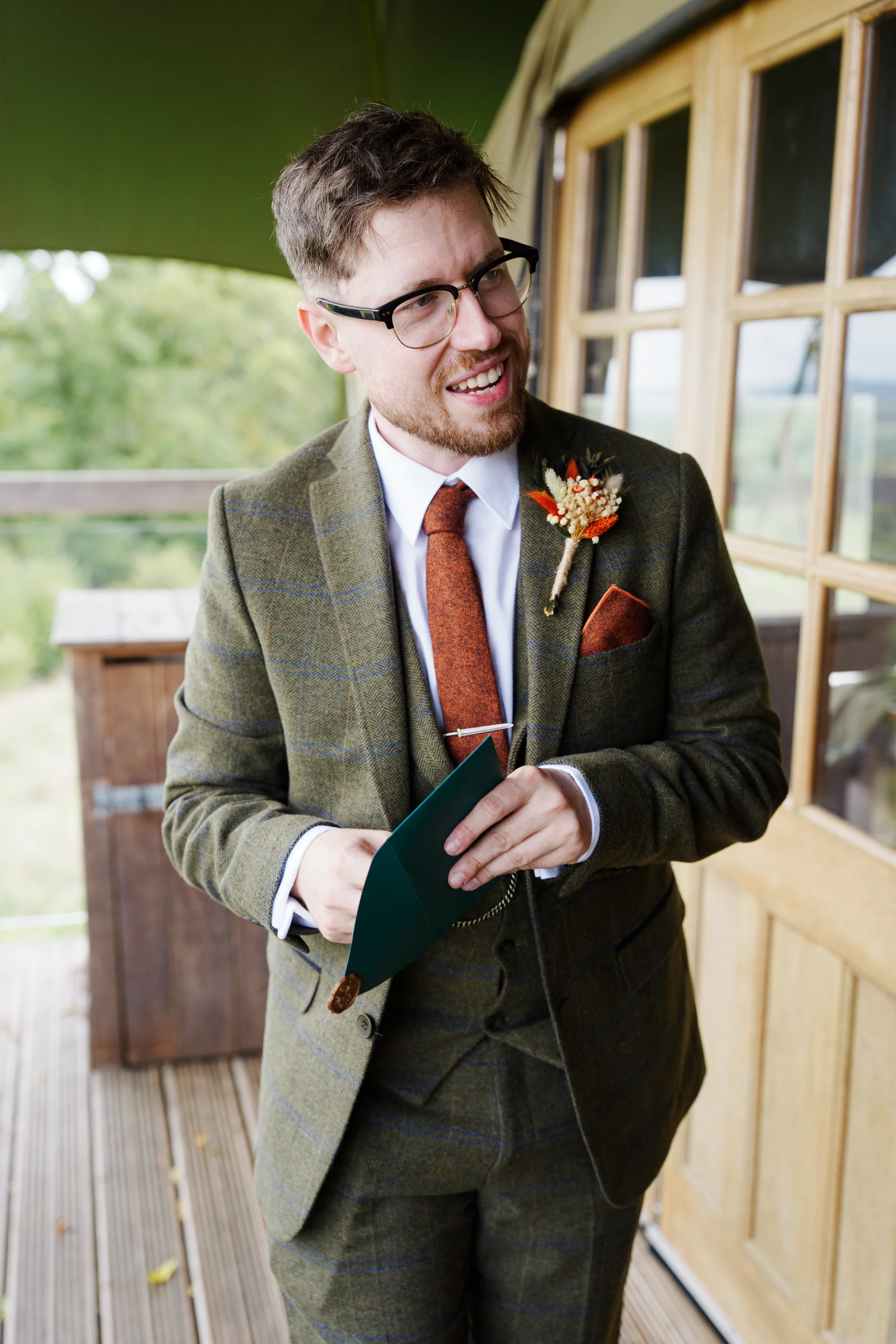 A smiling groom wearing a green tweed suit and a rust-coloured tie reads a sealed card while standing on a wooden deck.