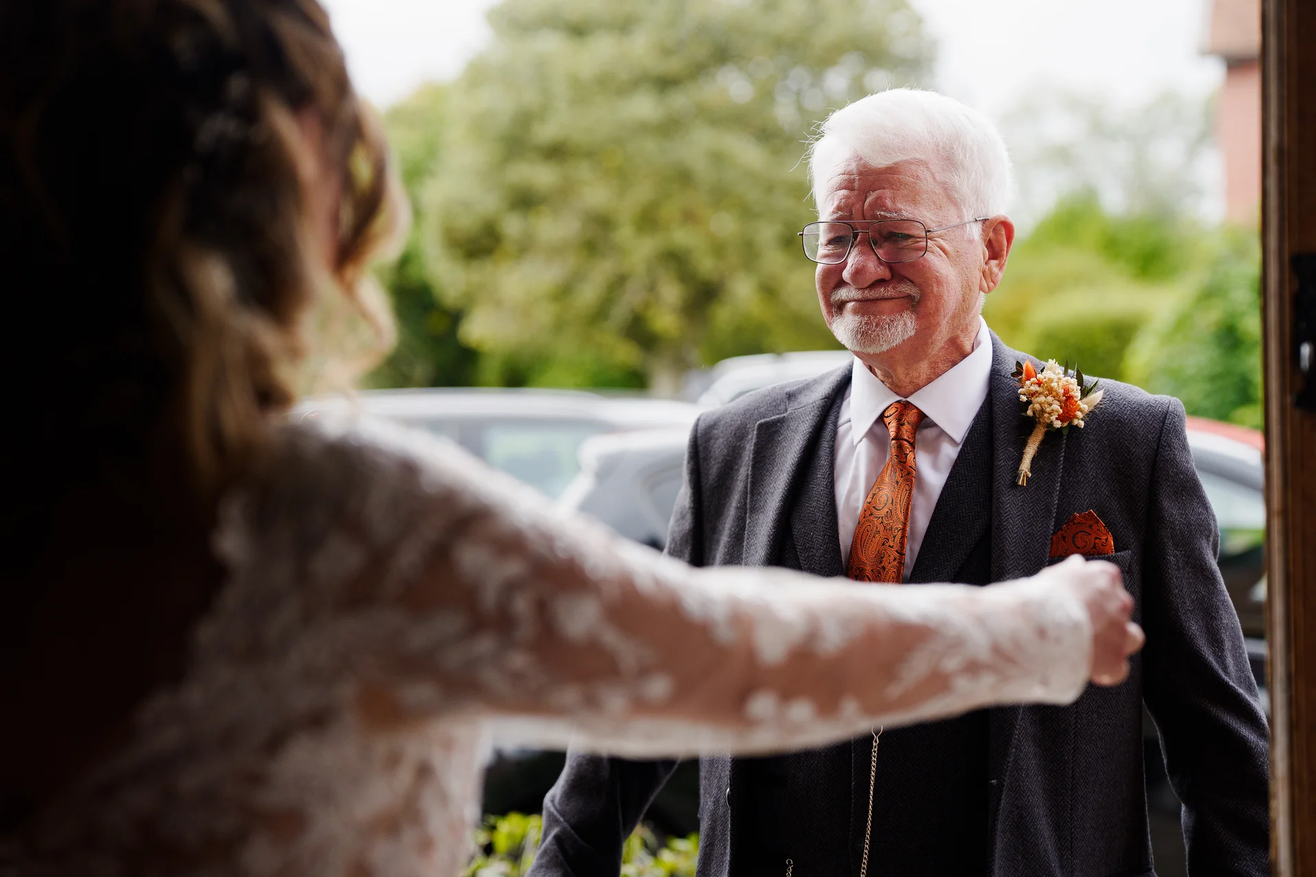An emotional father of the bride wearing a charcoal tweed suit and patterned tie looks proudly at his daughter in her lace wedding dress during a first look moment.