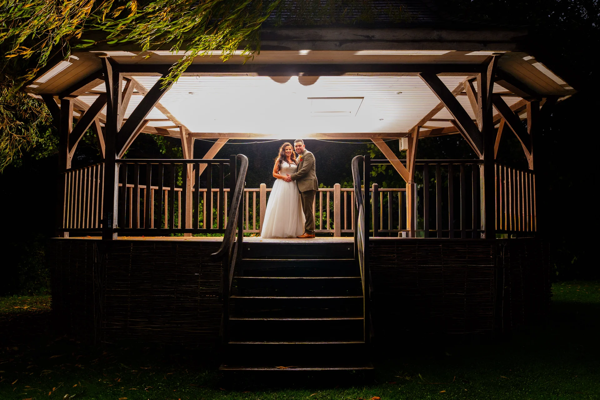 A newlywed couple stands embraced inside an illuminated wooden gazebo at night, framed by foliage and dramatic overhead lighting.
