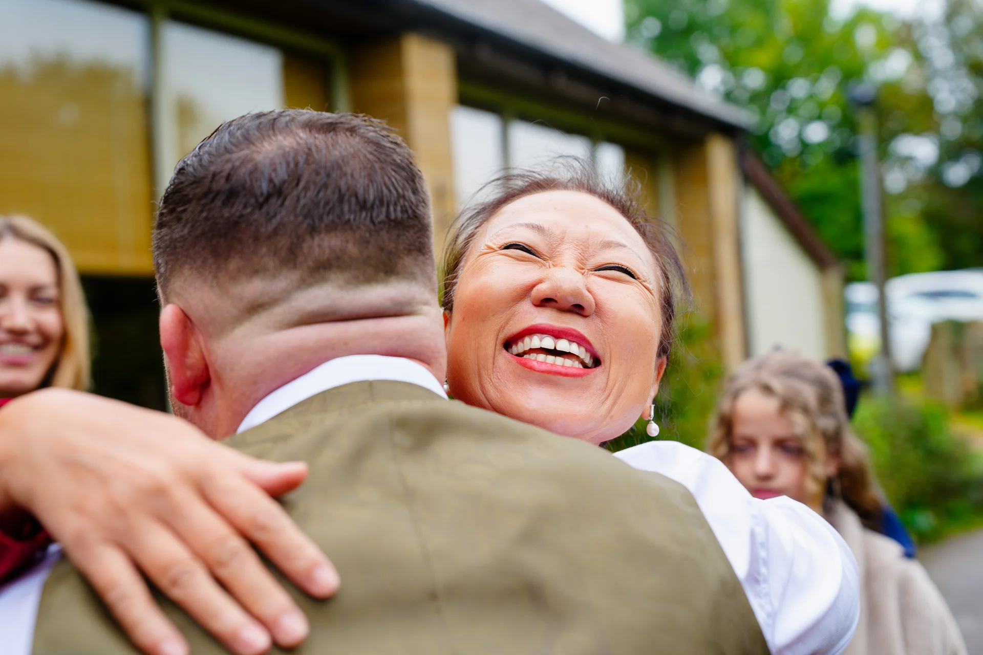 A mature woman embraces a groom tightly outside a venue, laughing joyfully with her eyes closed in a candid moment.