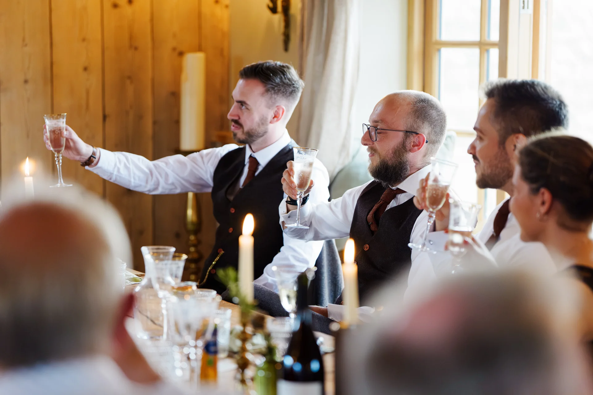 Groomsmen and guests raise champagne flutes in a toast during the wedding breakfast inside a rustic wood-panelled venue.