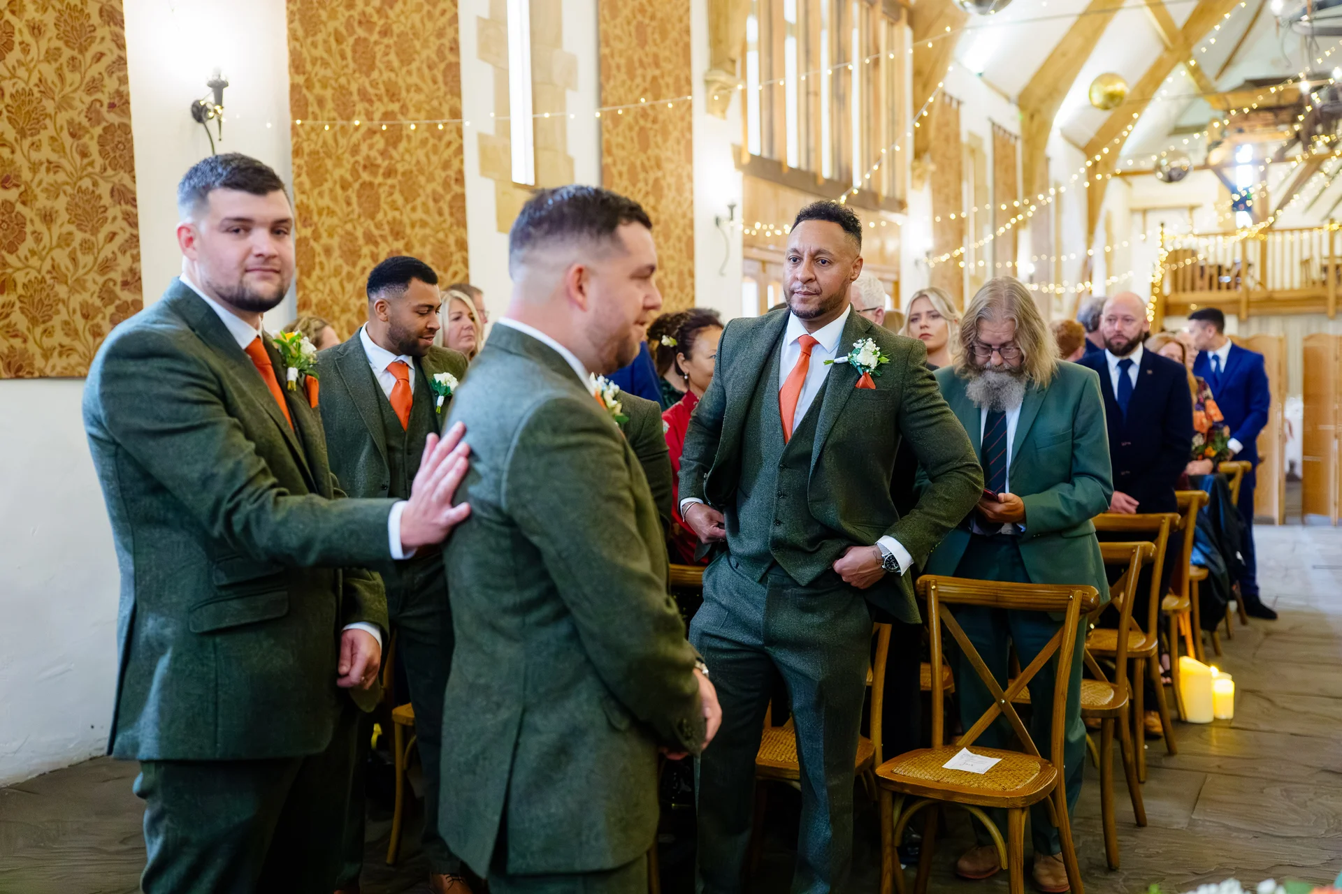 A group of groomsmen wearing matching green tweed suits and orange ties stand inside a rustic barn venue decorated with twinkling fairy lights.