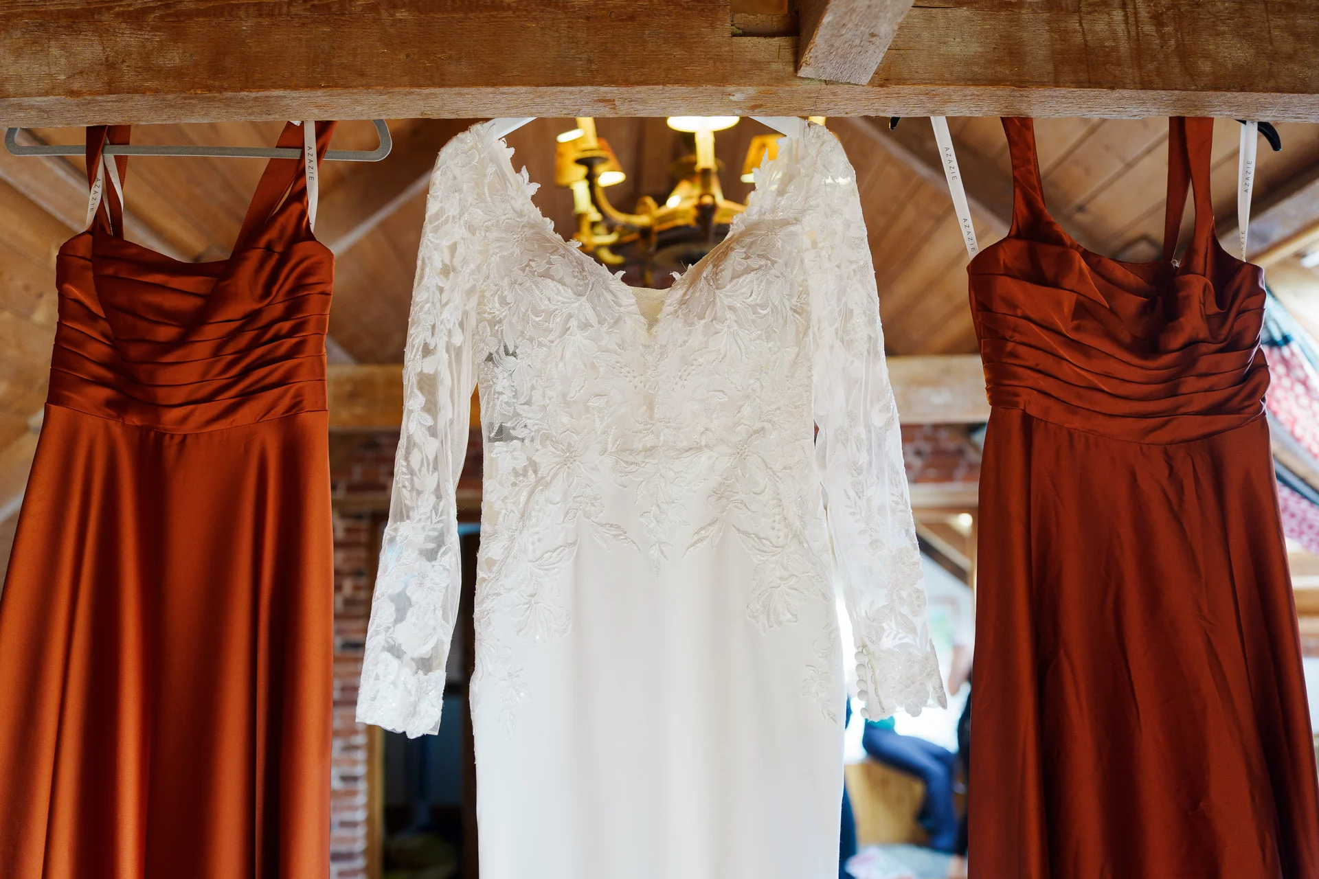 A white lace long-sleeved wedding dress hangs flanked by two rust-coloured bridesmaid dresses against a rustic wooden beam interior.