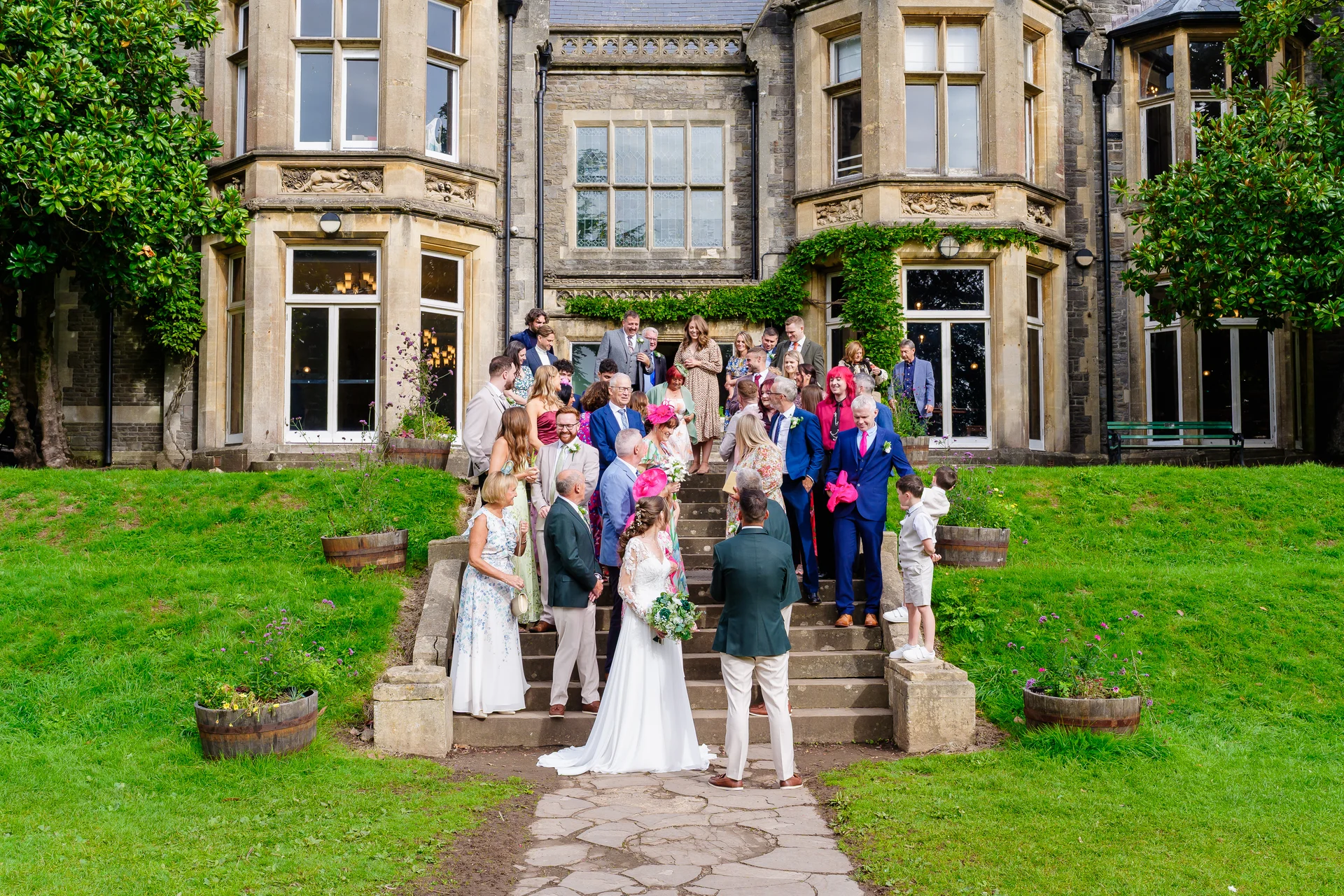 A newlywed couple and their wedding guests pose for a large group photograph on the grand stone steps outside a stately country manor house.