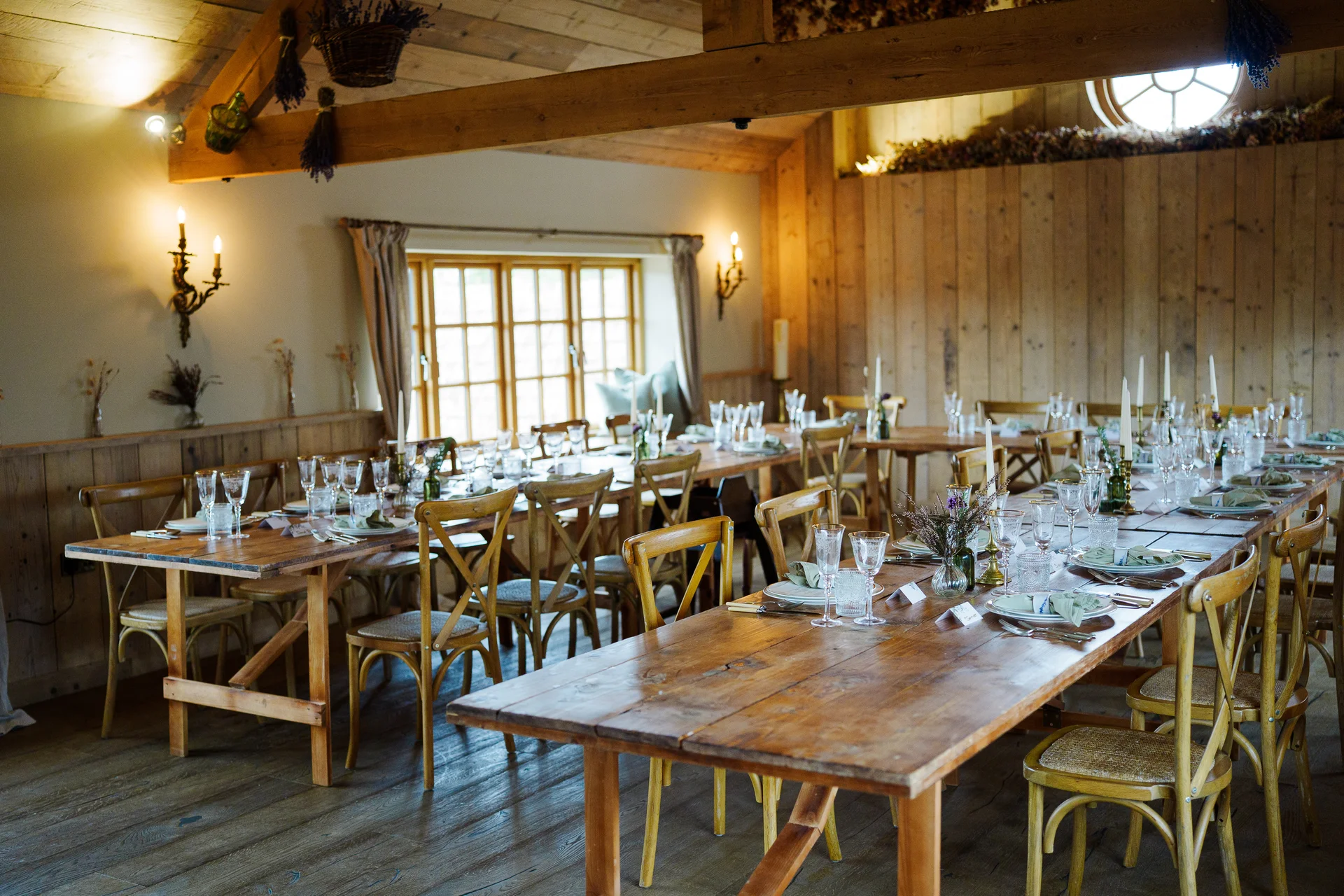 An elegantly laid rustic wooden banquet table inside a sunlit oak-framed barn wedding venue features delicate glassware and subtle natural decor.