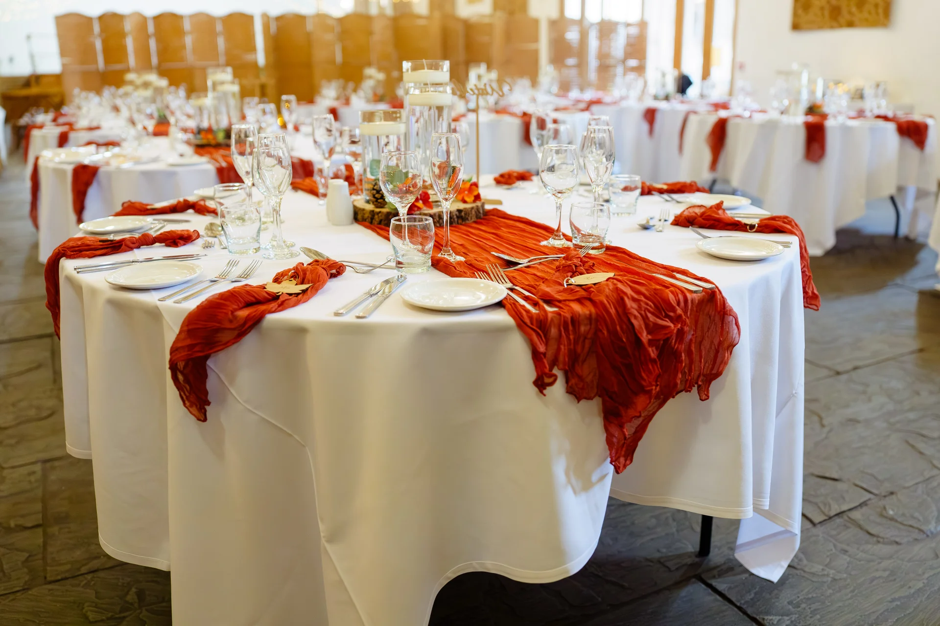 A beautifully styled wedding tablescape features white tablecloths draped with striking burnt orange runners, elegant glassware, and flickering candles.