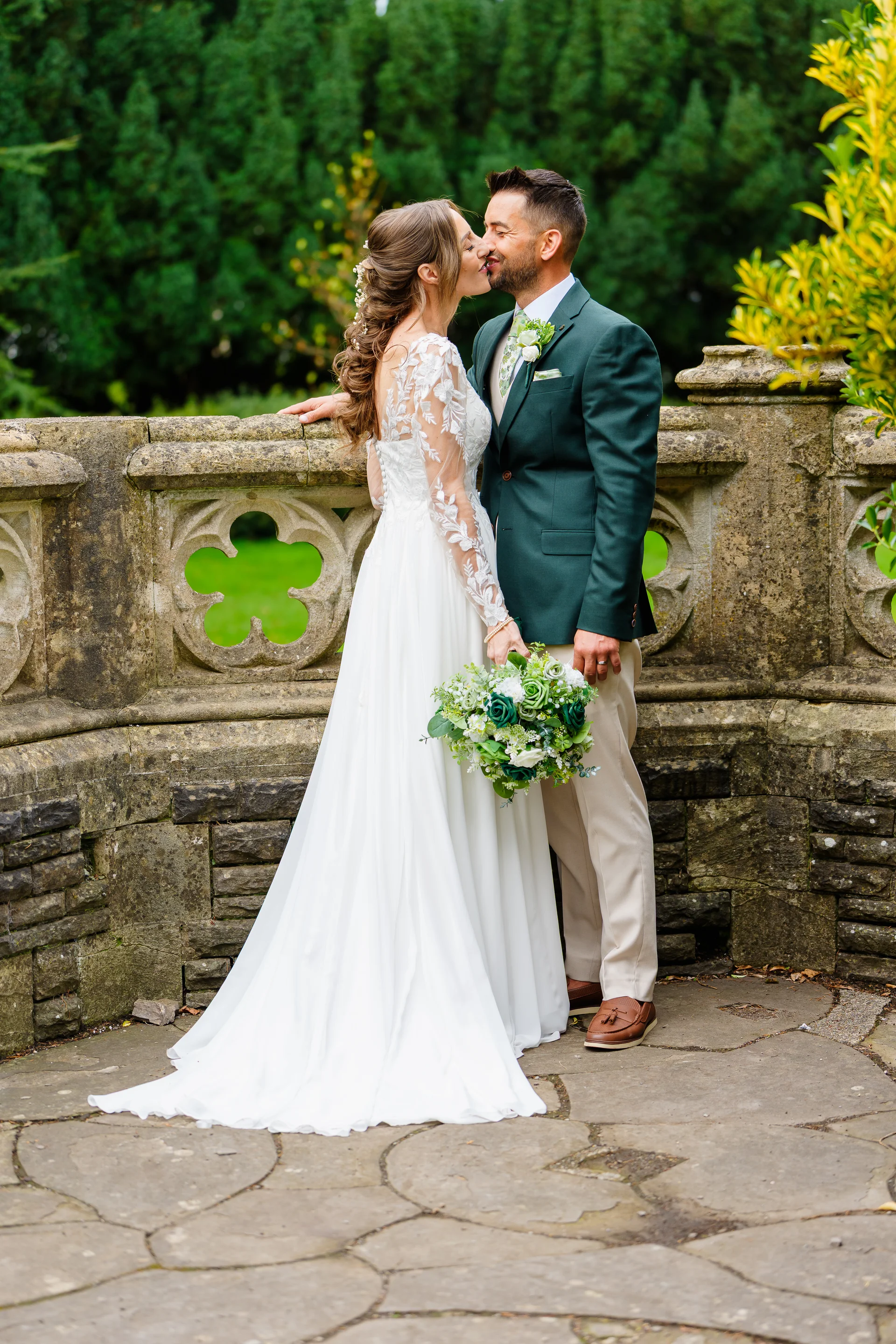 A bride and groom share a tender kiss outdoors next to an ornate stone balustrade in a lush garden setting.