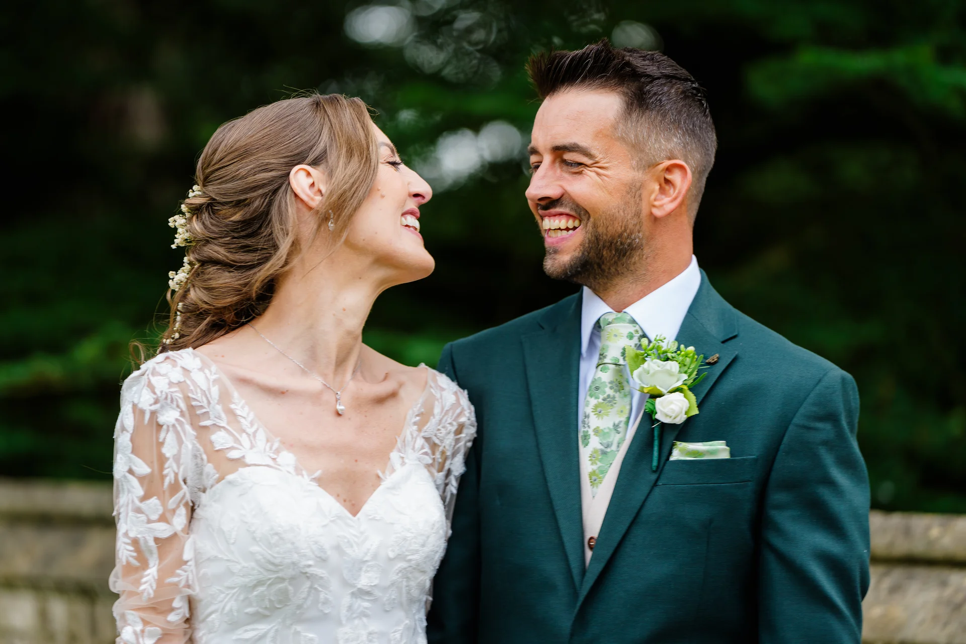 A newlywed bride and groom smile joyfully at each other during an outdoor portrait session, with the bride wearing a lace gown and the groom in a smart green suit.