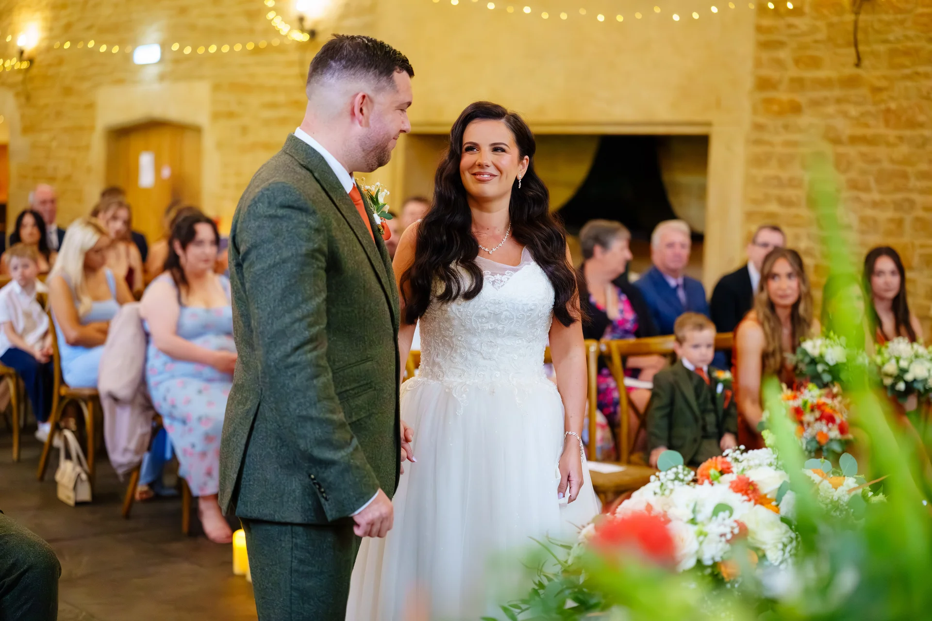 A smiling bride and groom hold hands during their indoor ceremony inside a rustic stone barn venue surrounded by seated guests and floral arrangements.