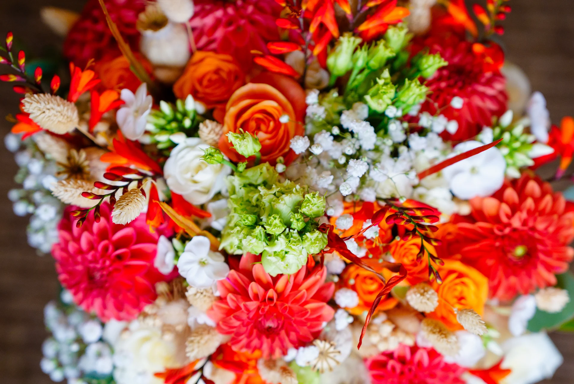 A vibrant overhead close-up shot of a richly textured wedding bouquet featuring an array of bright red dahlias, striking orange roses, and delicate white baby's breath.