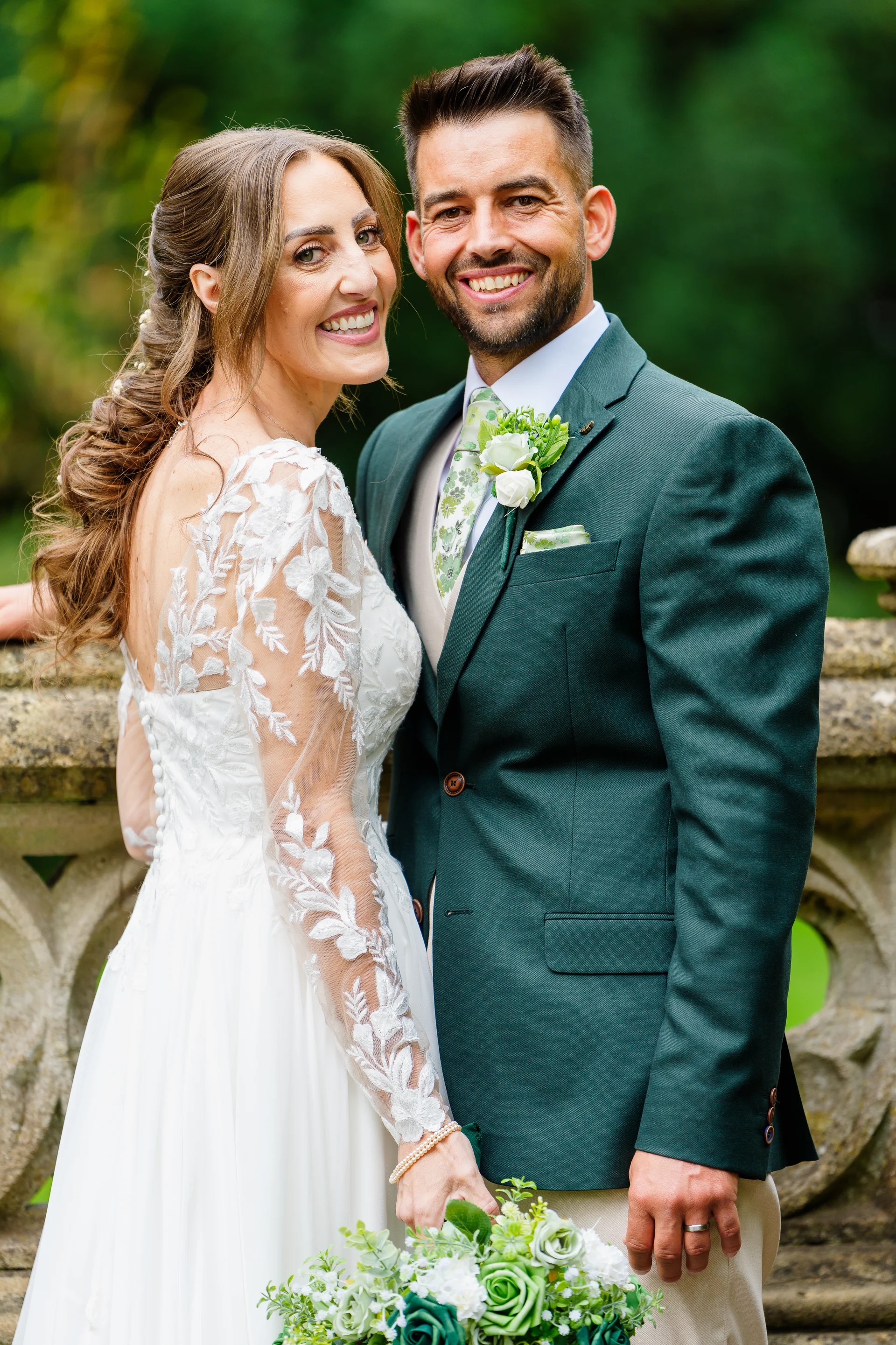 A smiling bride and groom pose together outdoors near a stone balustrade, with the bride wearing a lace-sleeved gown and the groom in a dark green suit.