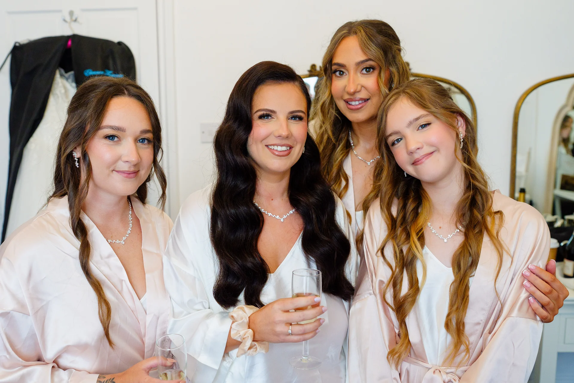 Four smiling women wearing matching satin robes and delicate jewellery pose together during wedding morning preparations, holding champagne glasses.