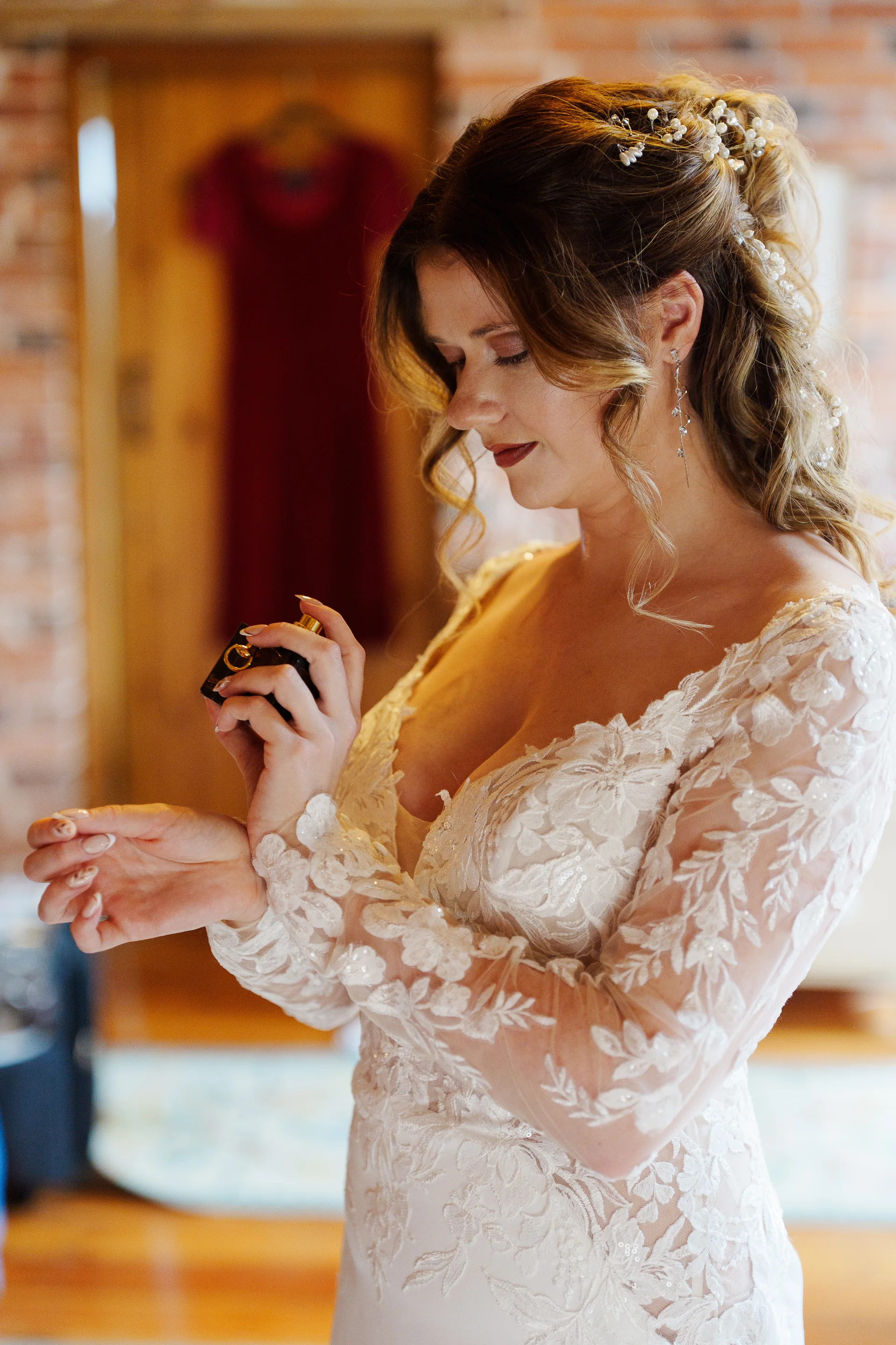A stunning bride delicately sprays perfume onto her wrist during bridal preparations in a room with exposed brick walls.