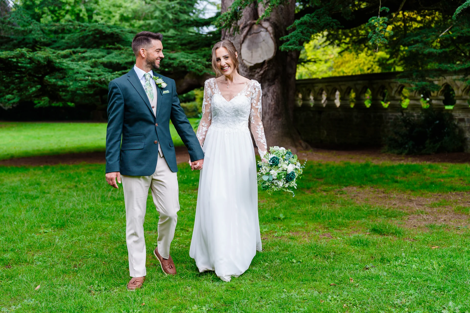 A newlywed couple happily walking hand in hand across a lush green lawn in front of an ancient tree and stone wall.