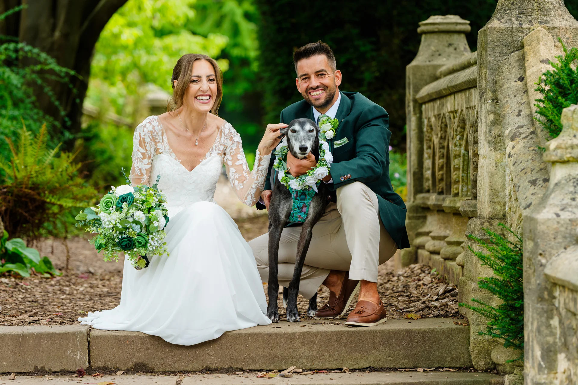 A smiling bride and groom sit happily on stone steps outdoors with their greyhound dog wearing a floral collar, while the bride holds a white and green bouquet.