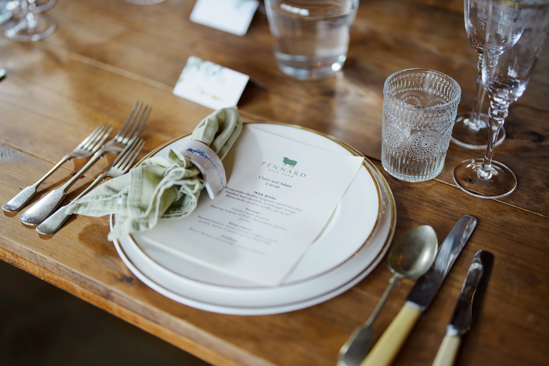 A beautifully styled rustic wedding tablescape featuring a wooden table, layered white plates with gold trim, a printed menu, and sage green linen napkins.