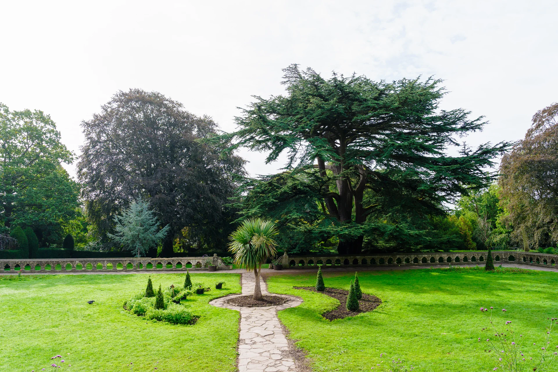 A wide angle view of the manicured grounds featuring a stone path leading through green lawns towards a grand ancient tree and an ornate stone balustrade.