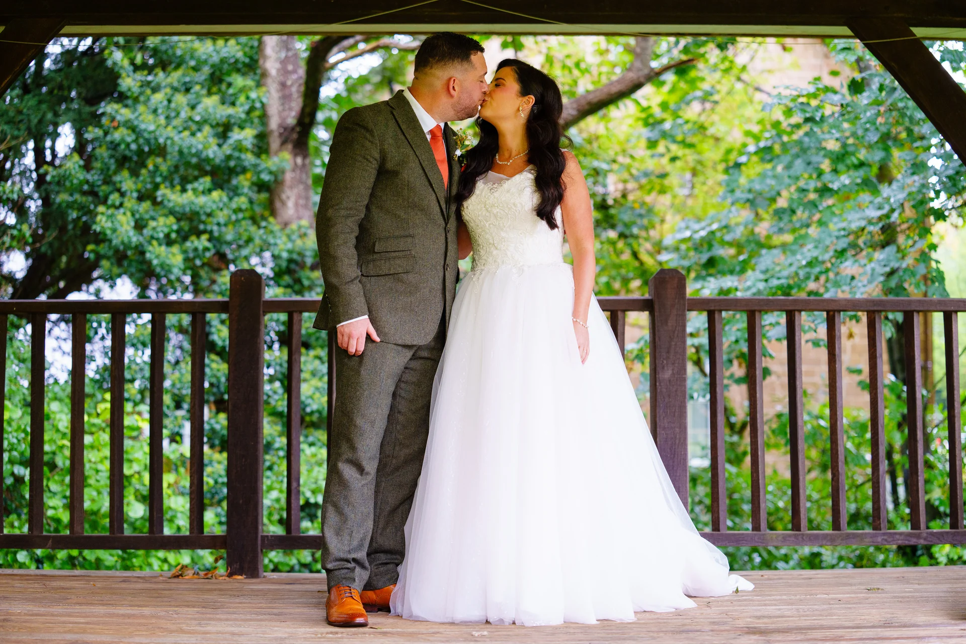 A newlywed couple shares a tender kiss inside a wooden gazebo surrounded by lush green trees on their wedding day.