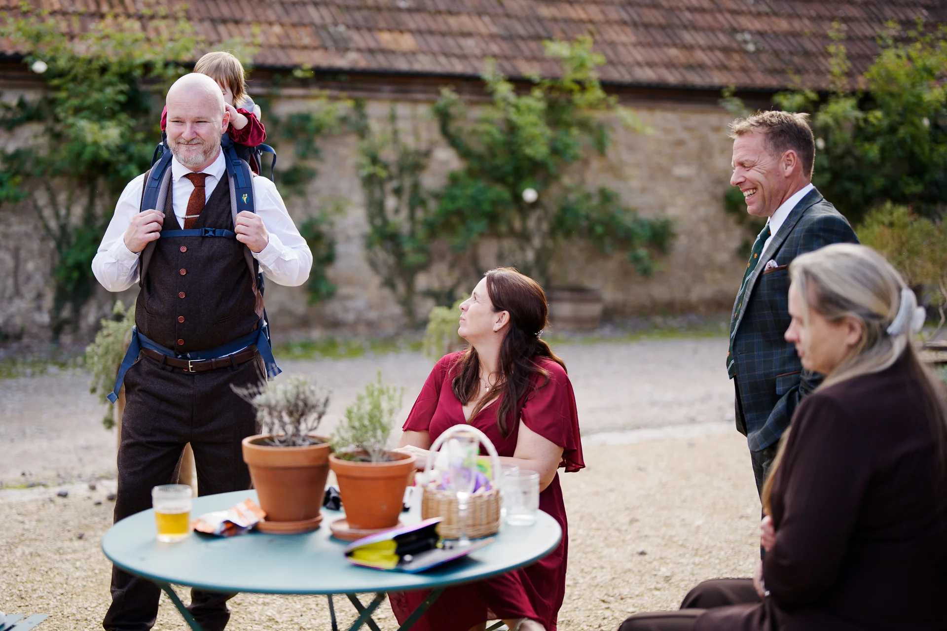Wedding guests laugh and socialise outdoors in a stone courtyard, with a man carrying a child on his shoulders near a small table with potted plants.