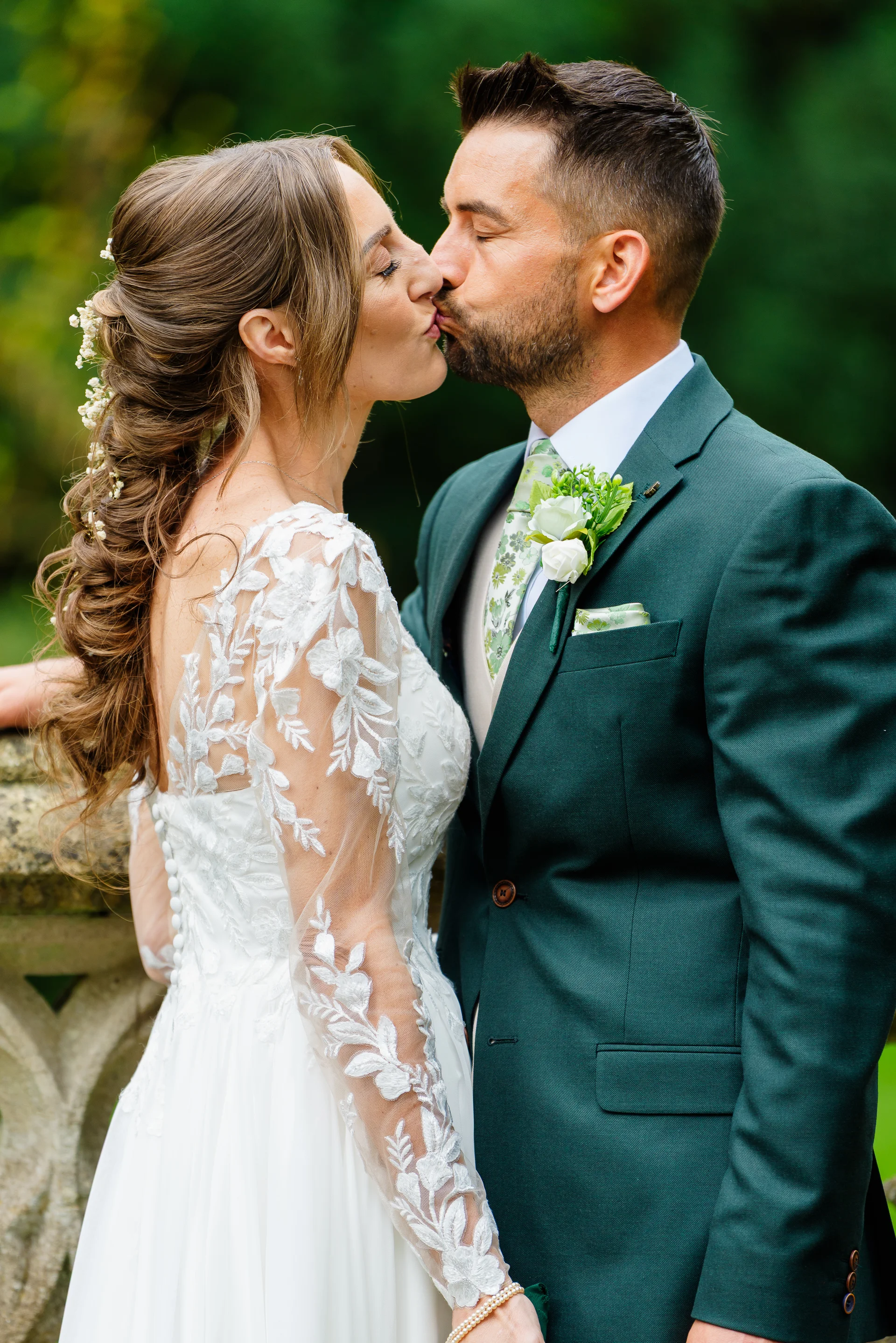 A newlywed couple shares an intimate kiss outdoors, with the bride wearing a long-sleeved lace gown and delicate flowers in her hair, and the groom in a smart green suit.