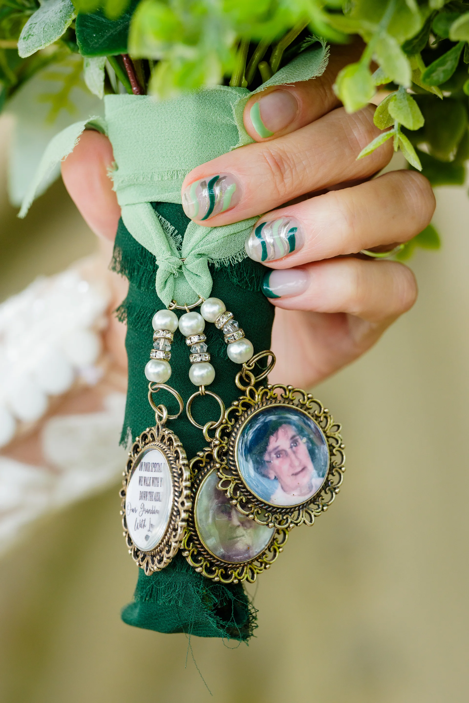 Close-up of a bridal hand holding a green ribbon-wrapped bouquet adorned with vintage-style photo charms of loved ones and custom green nail art.
