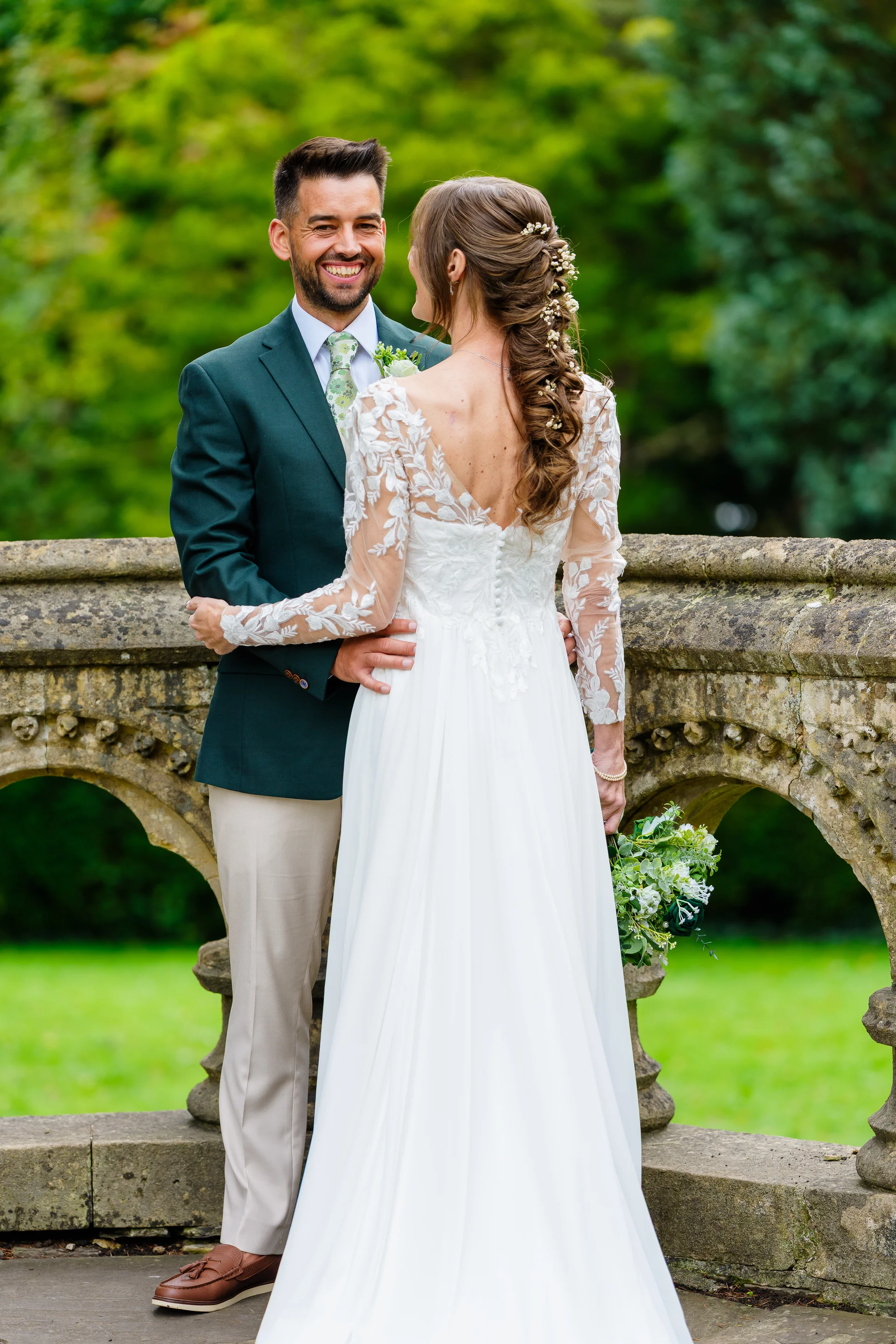A newlywed couple smiling at each other while embracing on a stone bridge outdoors, with the bride wearing a long-sleeve lace-detailed gown and the groom in a dark green suit.