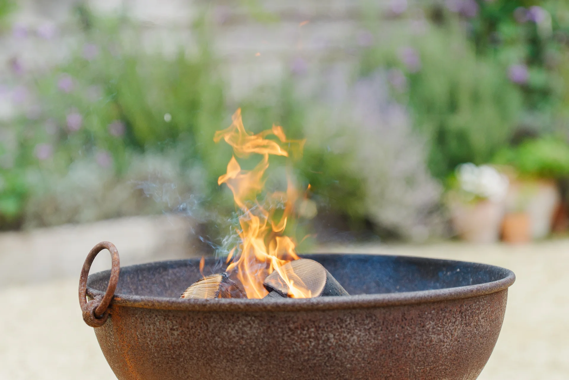 A rustic metal fire bowl filled with burning wood logs and gentle flames sits outdoors against a softly blurred green garden backdrop.
