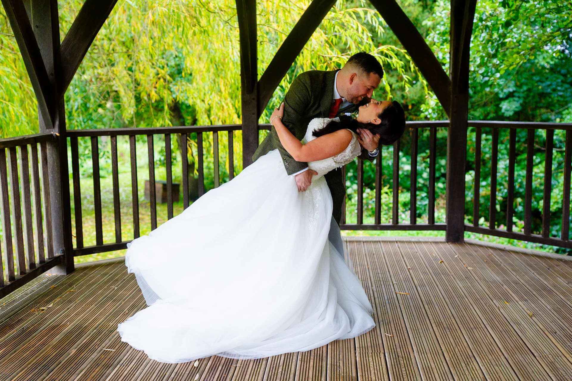 A groom dips his bride in a romantic kiss inside a wooden gazebo framed by lush green trees and foliage.