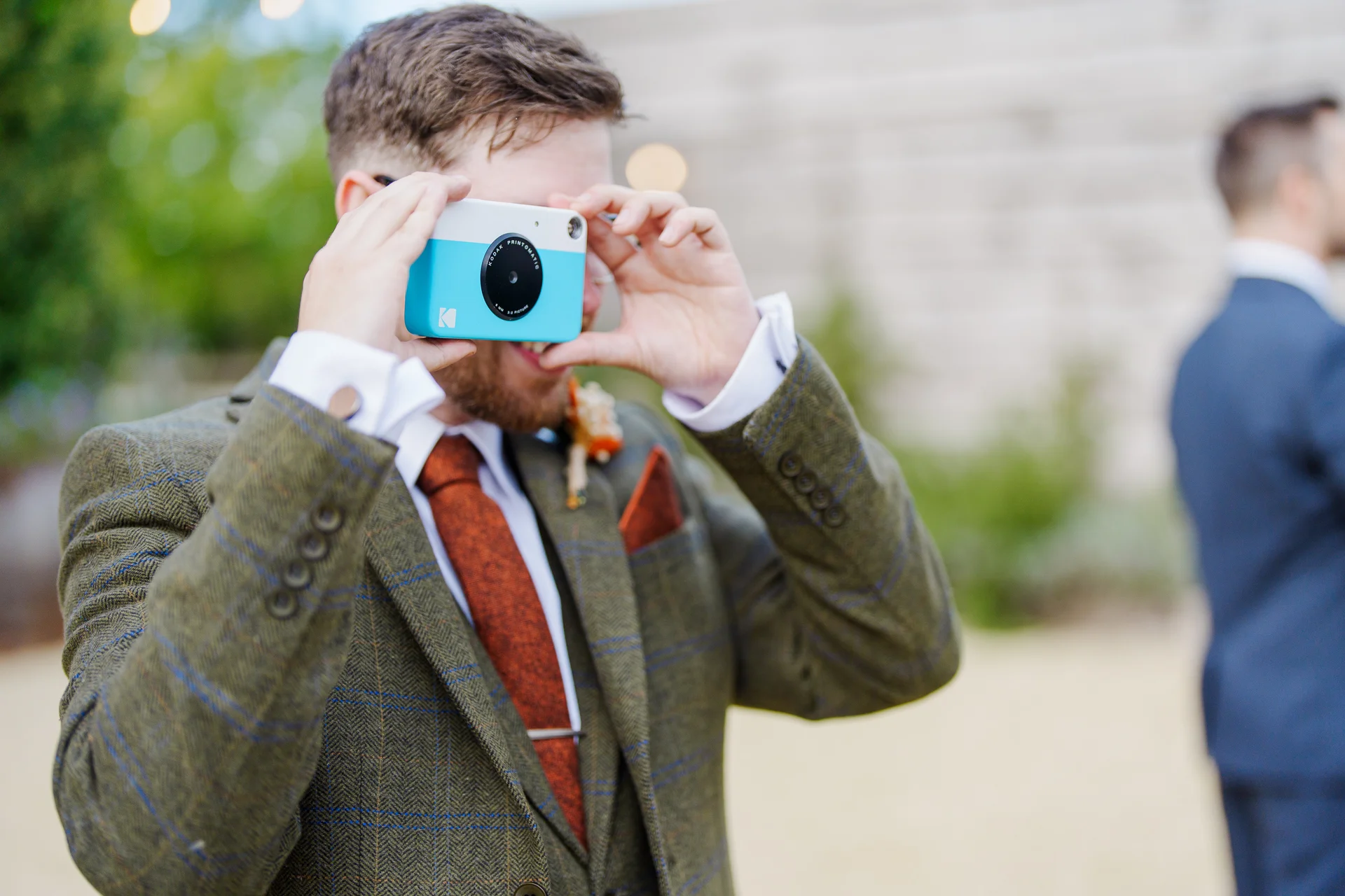 A groom wearing a green tweed suit and rust-coloured tie smiles as he holds a blue and white instant camera up to his face outdoors.