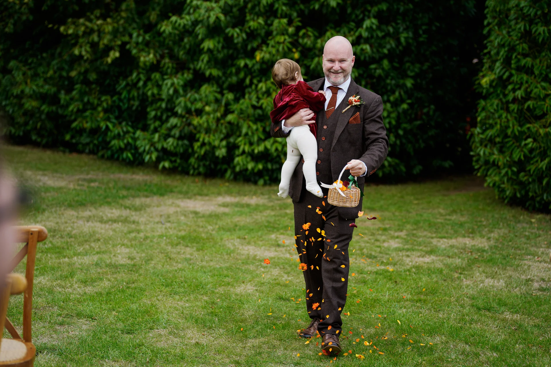 A smiling groom wearing a brown tweed suit carries a small child while walking across a lawn and scattering petals from a basket.