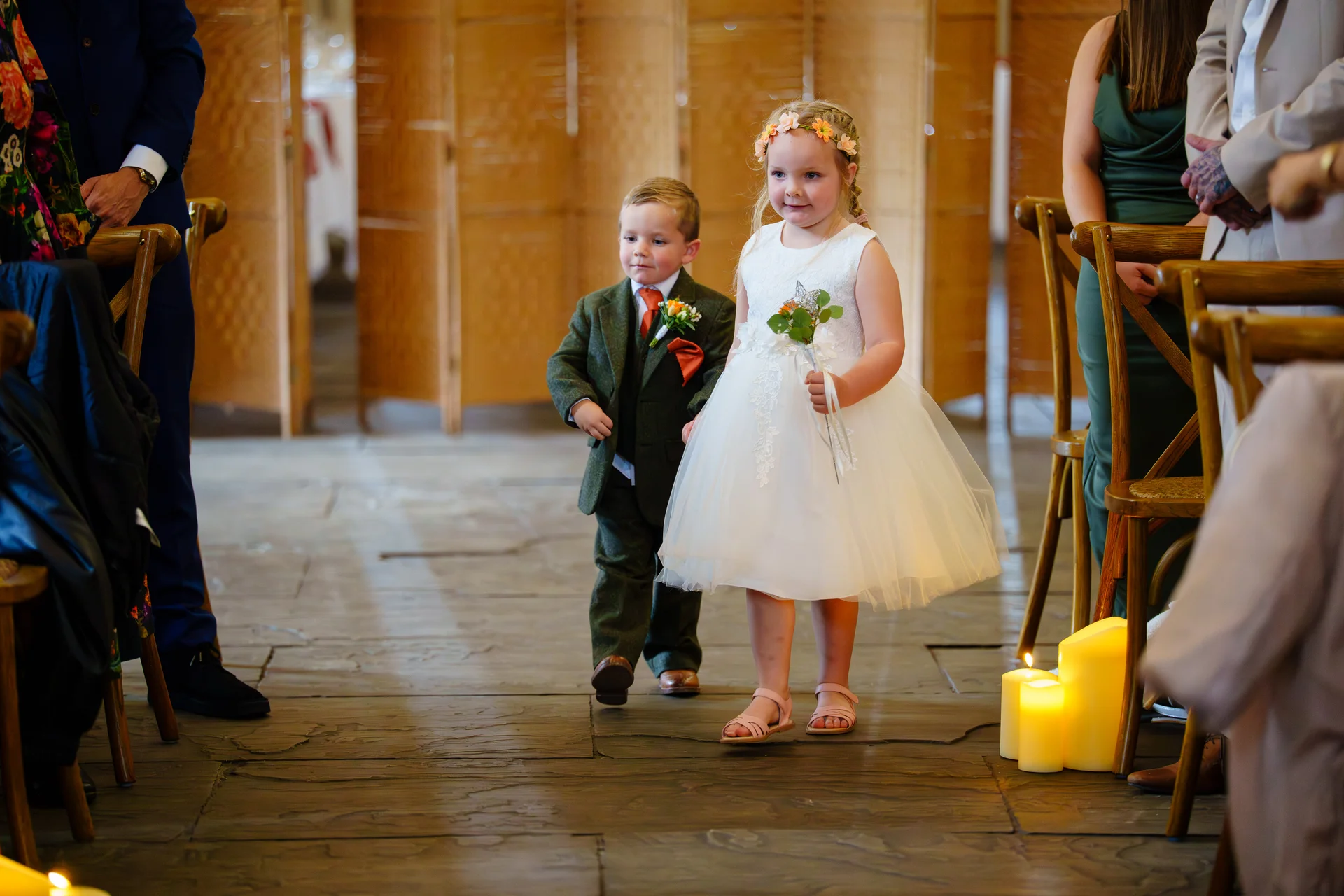 A young flower girl and page boy walk down the aisle together during a wedding ceremony, holding small floral arrangements.