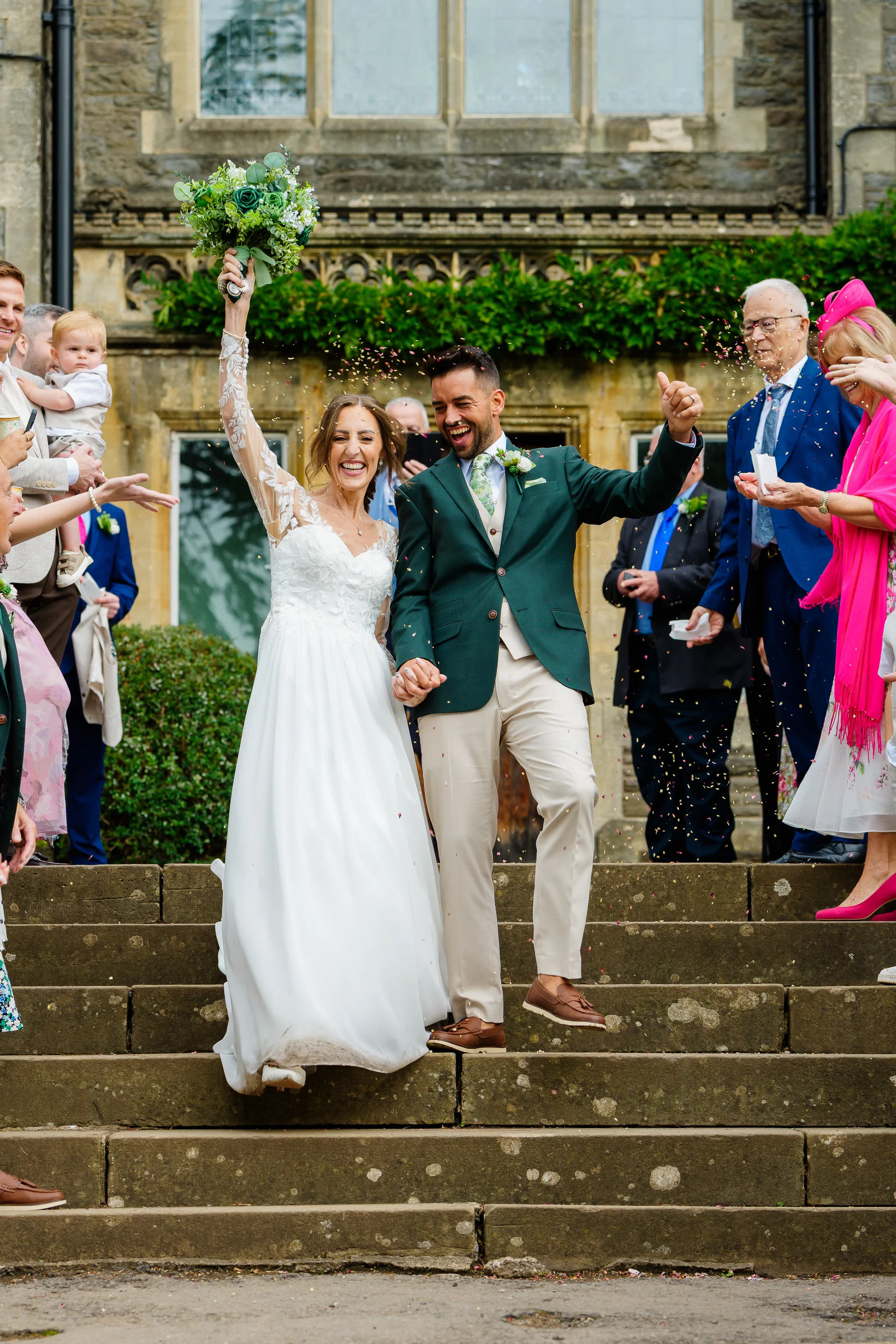 A joyful bride and groom walk down stone steps while guests throw confetti, celebrating their wedding in front of a historic country house.