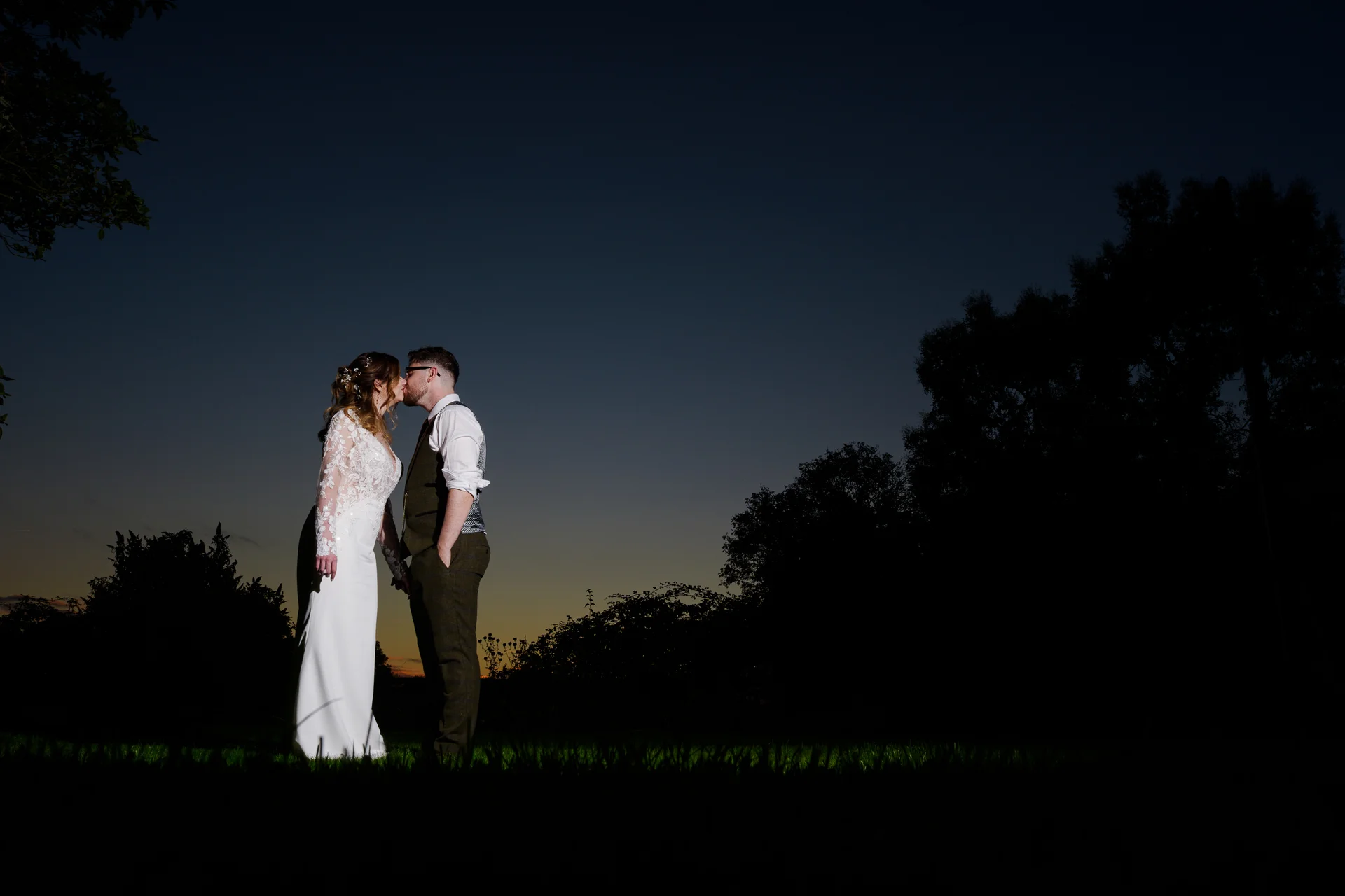 A newlywed couple shares a tender kiss outdoors at dusk against a dramatic dark sky and illuminated tree line.