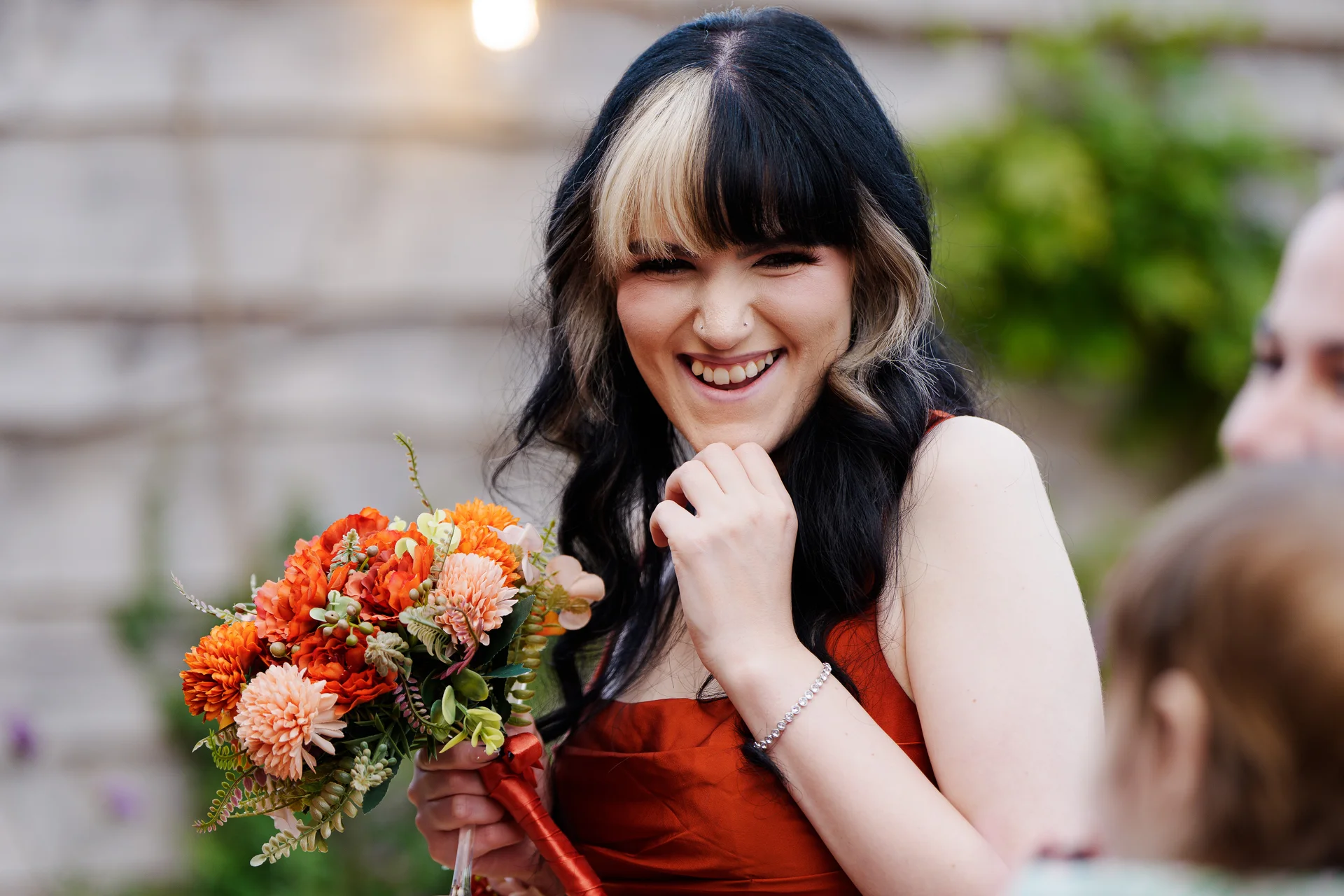 A smiling bride with dark hair and blonde highlights holds a vibrant orange bouquet of autumn flowers, laughing joyfully during her wedding.