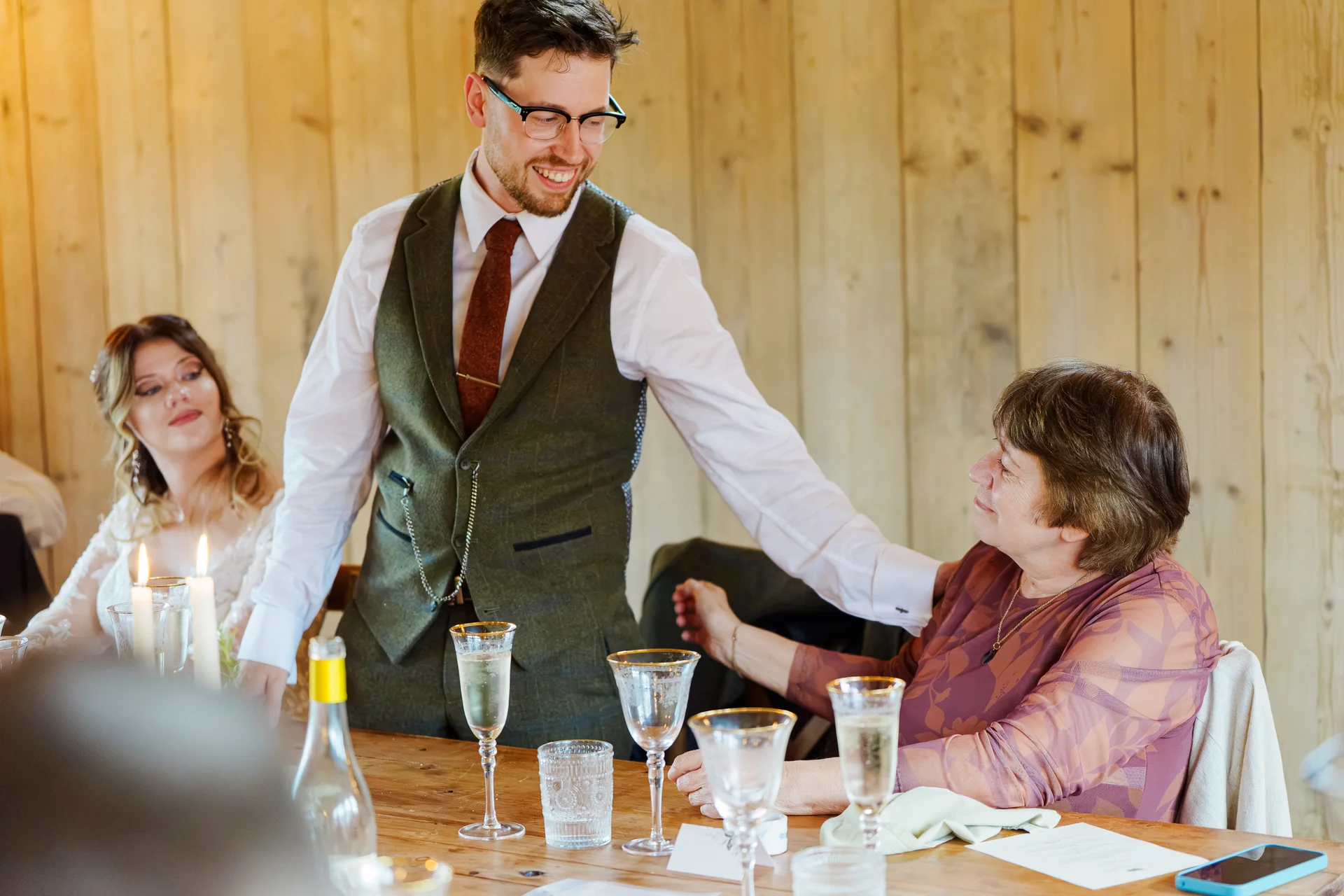 A smiling groom wearing a green tweed waistcoat leans in to speak with an older seated guest during the wedding reception speeches inside a rustic wooden barn.